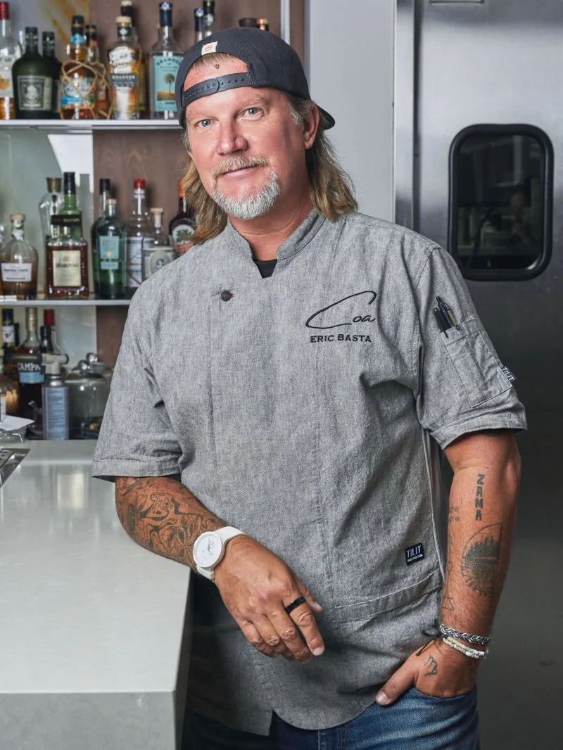 A man in a gray chef's coat with his name, Eric Basta, embroidered on it, standing in a bar or kitchen with liquor bottles on shelves behind him. He has tattoos on both arms, a white watch on his left wrist, and is wearing a backward black baseball cap.