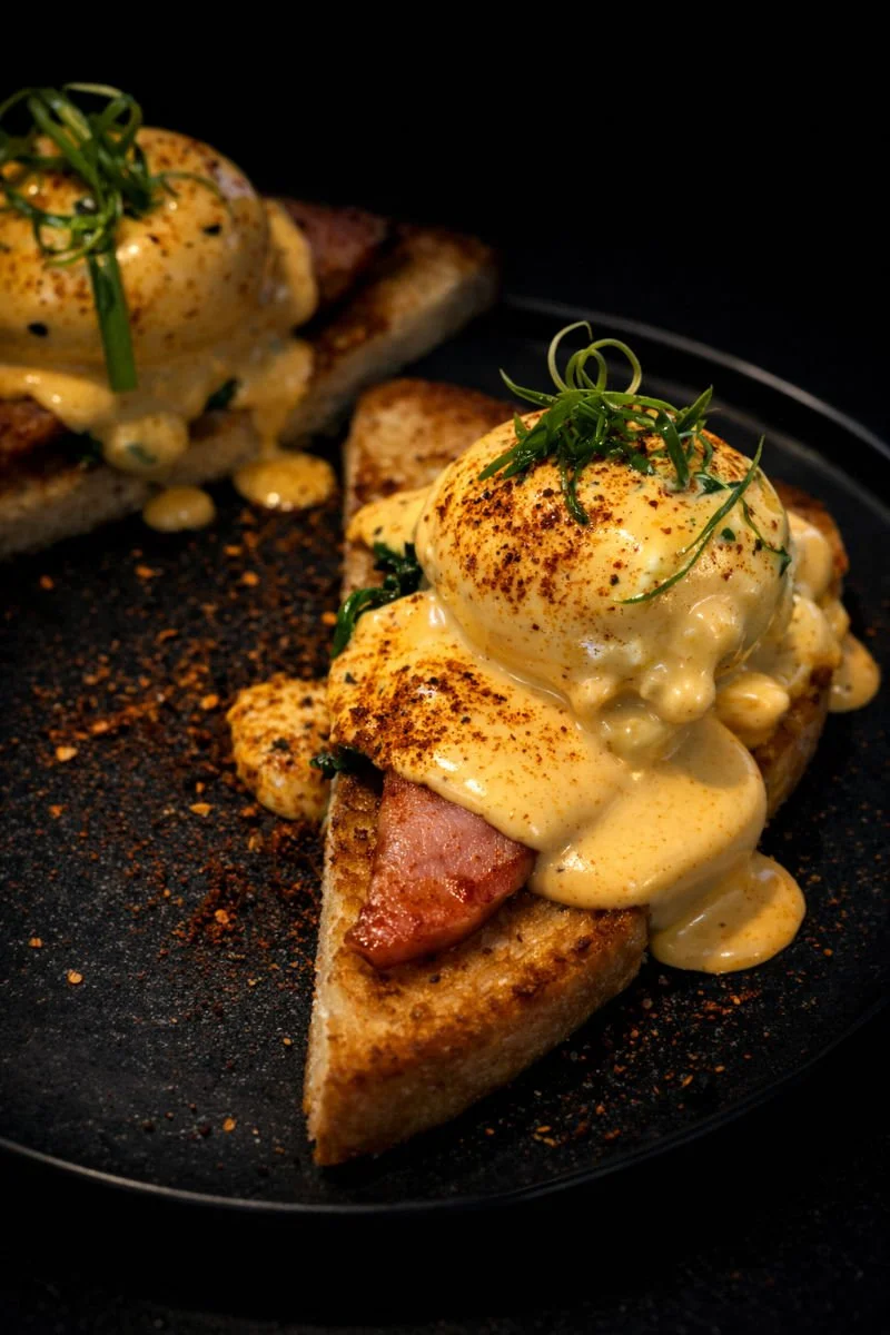 Eggs Benedict with poached eggs, hollandaise sauce, and green garnish on toasted bread with bacon, served on a black plate.