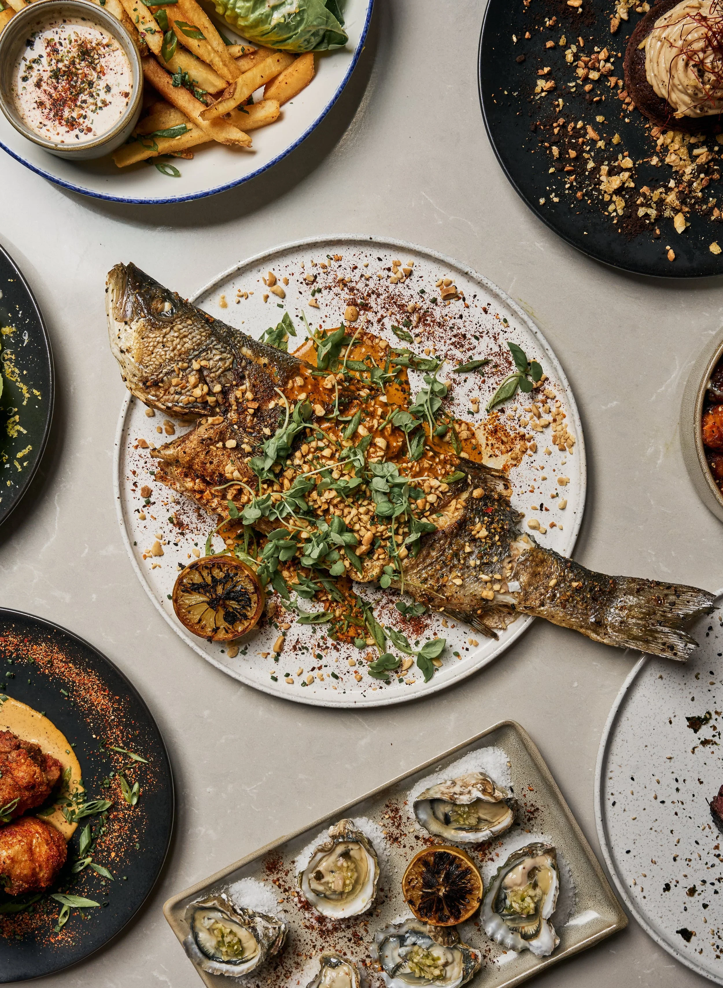 A table with various dishes, including a whole grilled fish topped with herbs and crushed nuts, served on a white plate. Surrounding dishes include oysters on a rectangular platter, grilled lemon halves, a bowl of fries with dipping sauce, and other assorted plates of food.