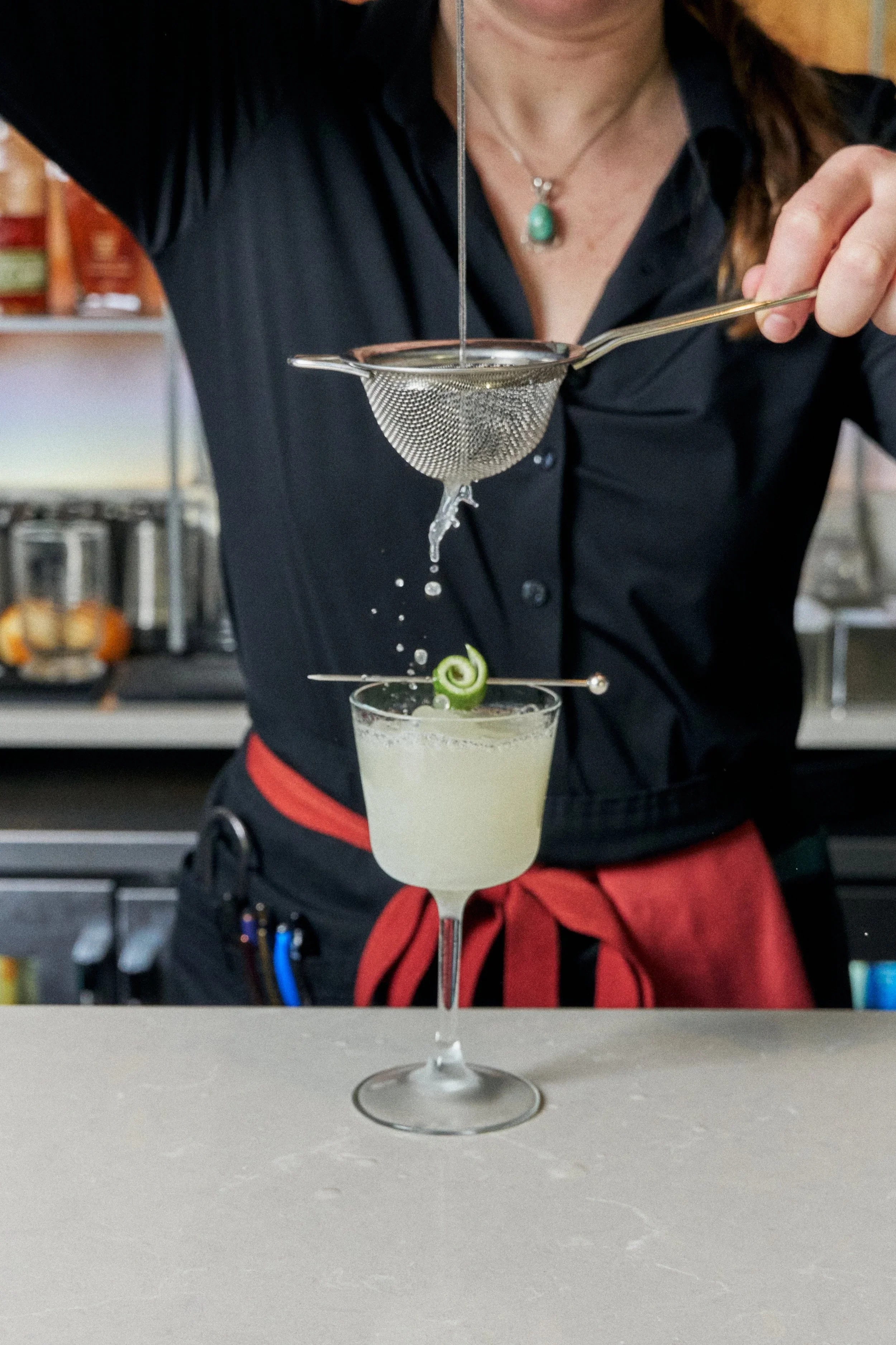 Bartender straining a cocktail into a glass garnished with a spiral cucumber, with liquid dripping through a fine strainer.
