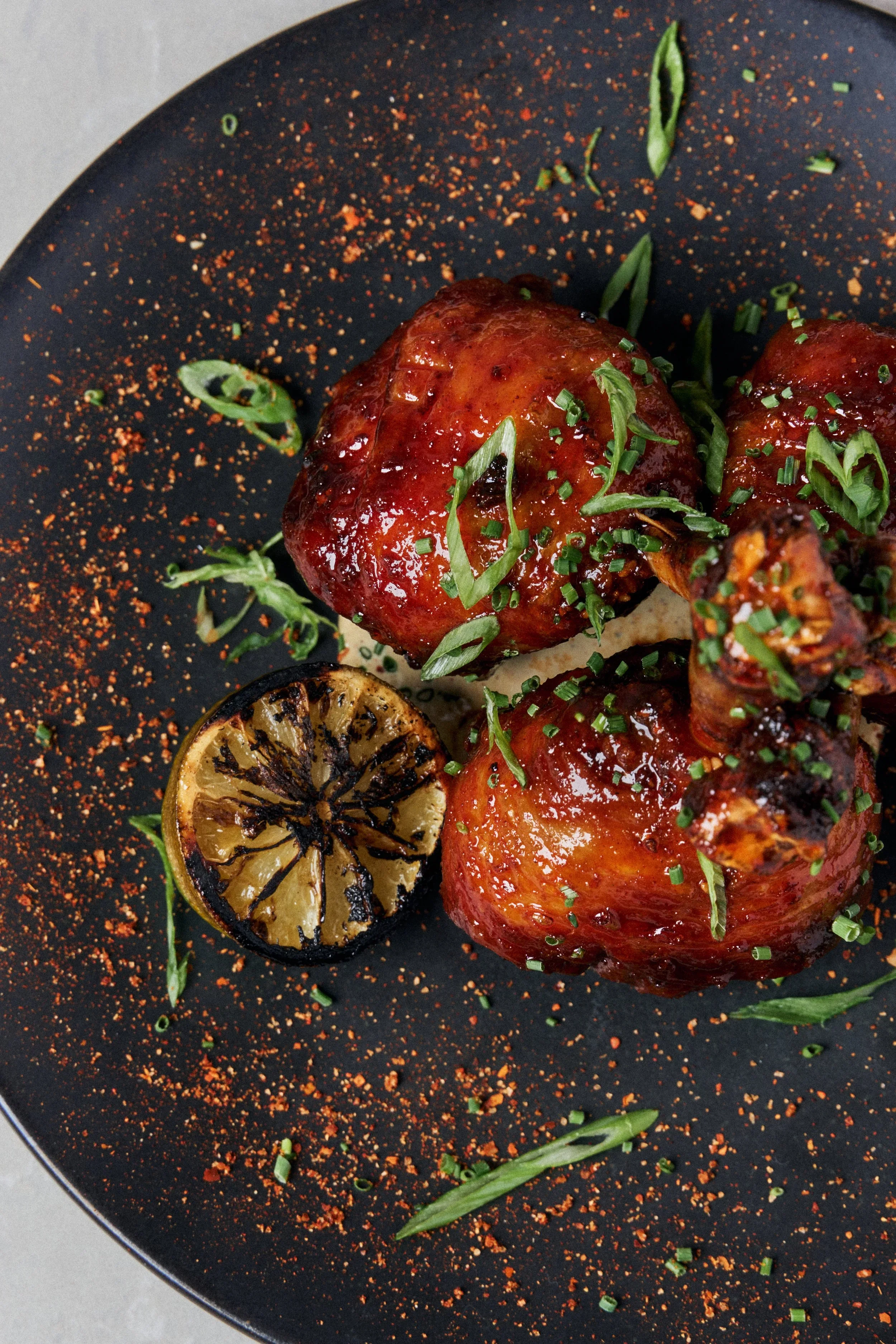 Close-up of glazed meatballs garnished with chopped green herbs, accompanied by a charred lemon half, on a black plate with red spice powder.