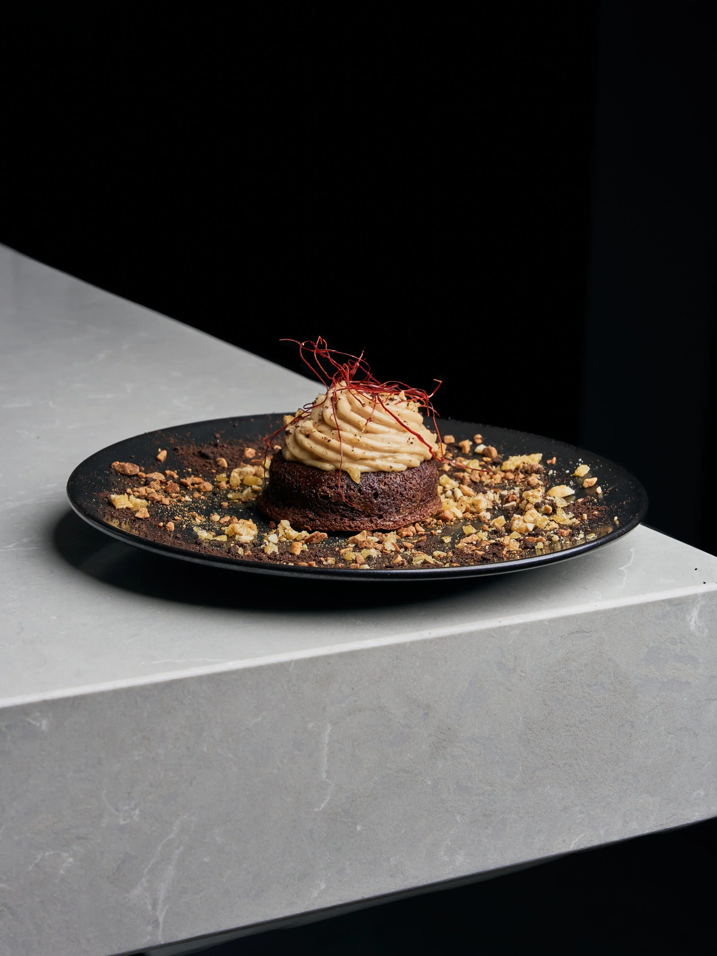 Chocolate dessert with cream topping, garnished with red threads, placed on a black plate with crumbled toppings, set on a white marble surface against a black background.