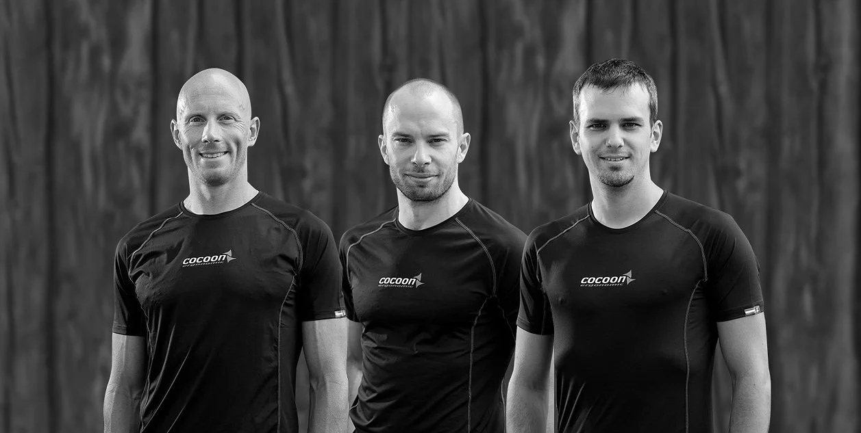 Drei Männer tragen schwarze Sportshirts mit dem Logo "cocoon" auf einem dunklen Holz-Hintergrund.