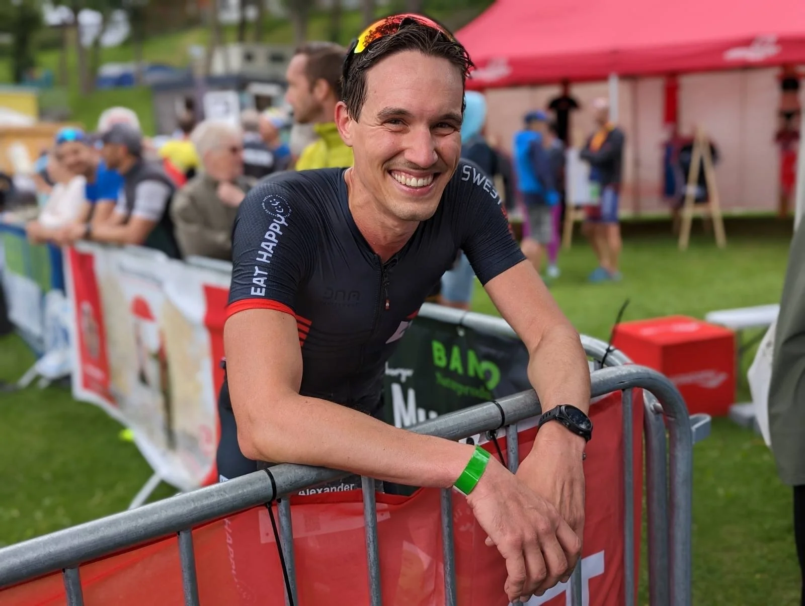 Lächelnder männlicher Triathlet an der Zielinie bei einem Wettkampf, umgeben von Zuschauern und einem Radherzungsbereich, trägt ein schwarzes Triathlon-Outfit mit roten und blauen Elementen.