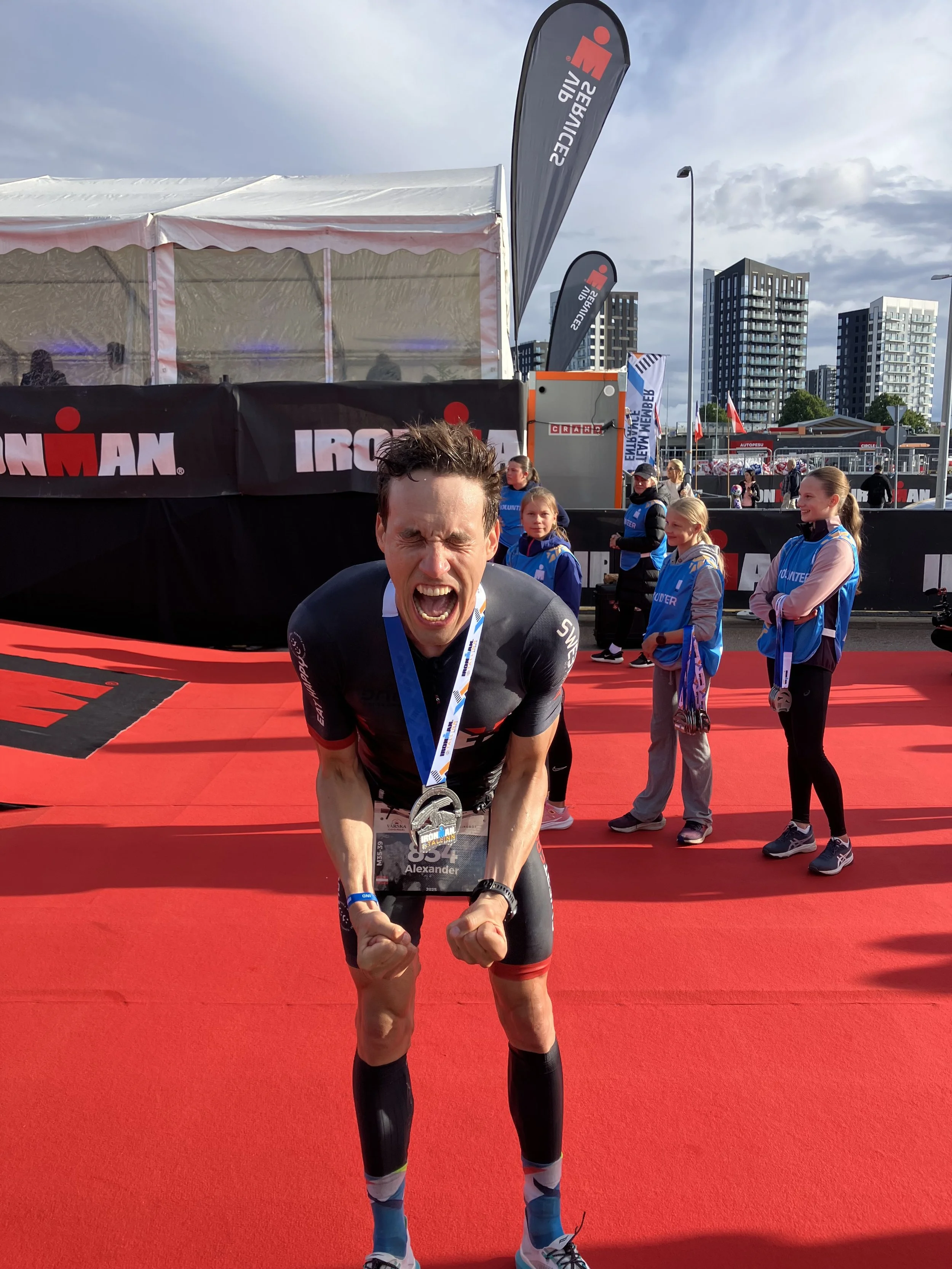 Ein Mann beim Ironman-Wettbewerb nach dem Ziel, mit Medaille um den Hals und Siegerpose, umgeben von Helfern und Zuschauern, im Stadtgebiet mit modernen Hochhäusern im Hintergrund.