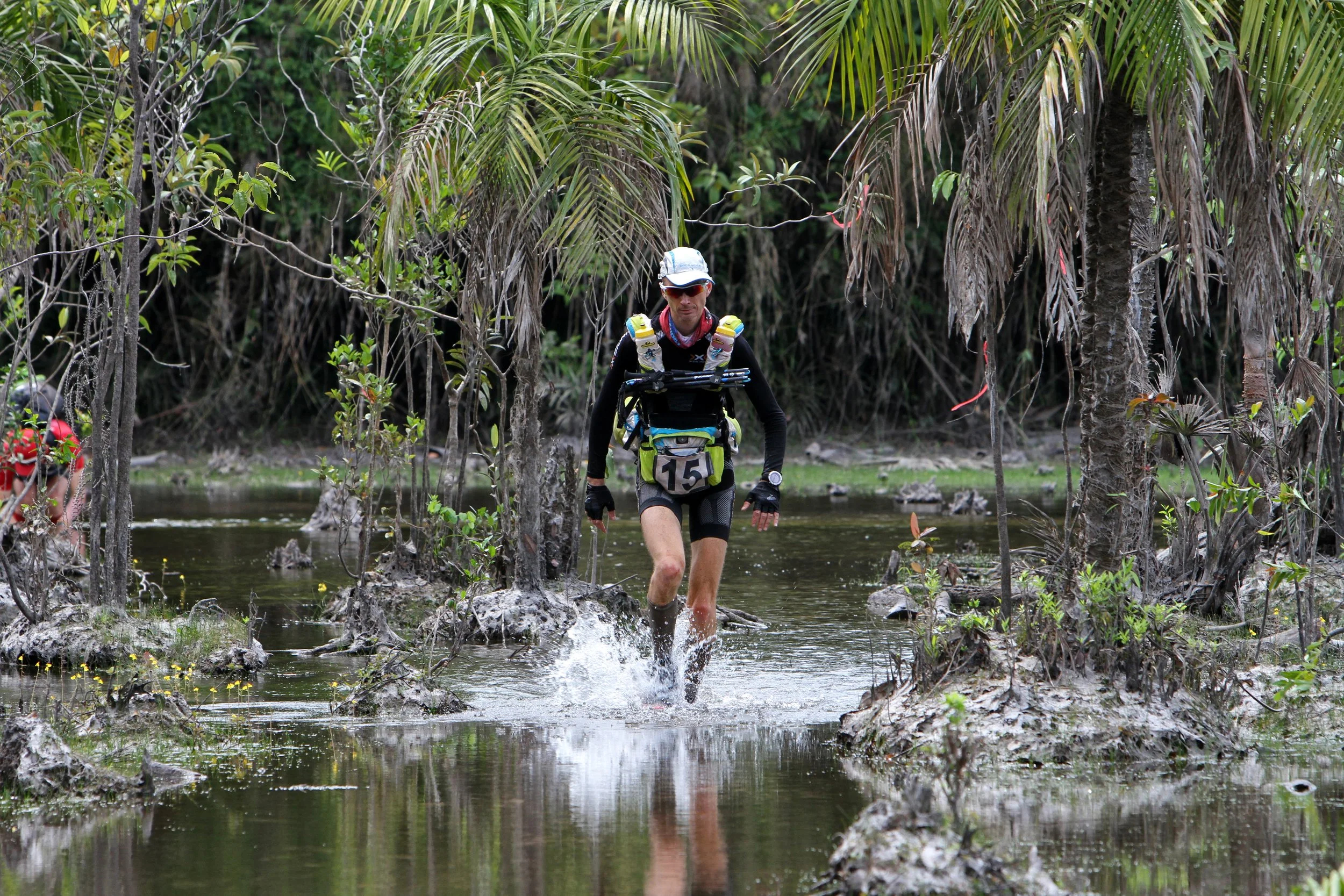 Ein Mountainbike-Fahrer in einem Wasserloch im Dschungel trägt Helm, Sonnenbrille und Rucksack.