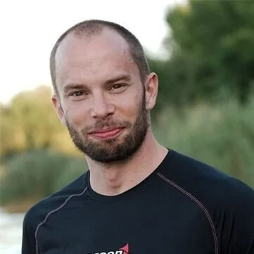 Ein Mann mit kurz geschorenem Haar und Bart, trägt ein schwarzes T-Shirt, steht im Freien mit grüner Natur im Hintergrund.