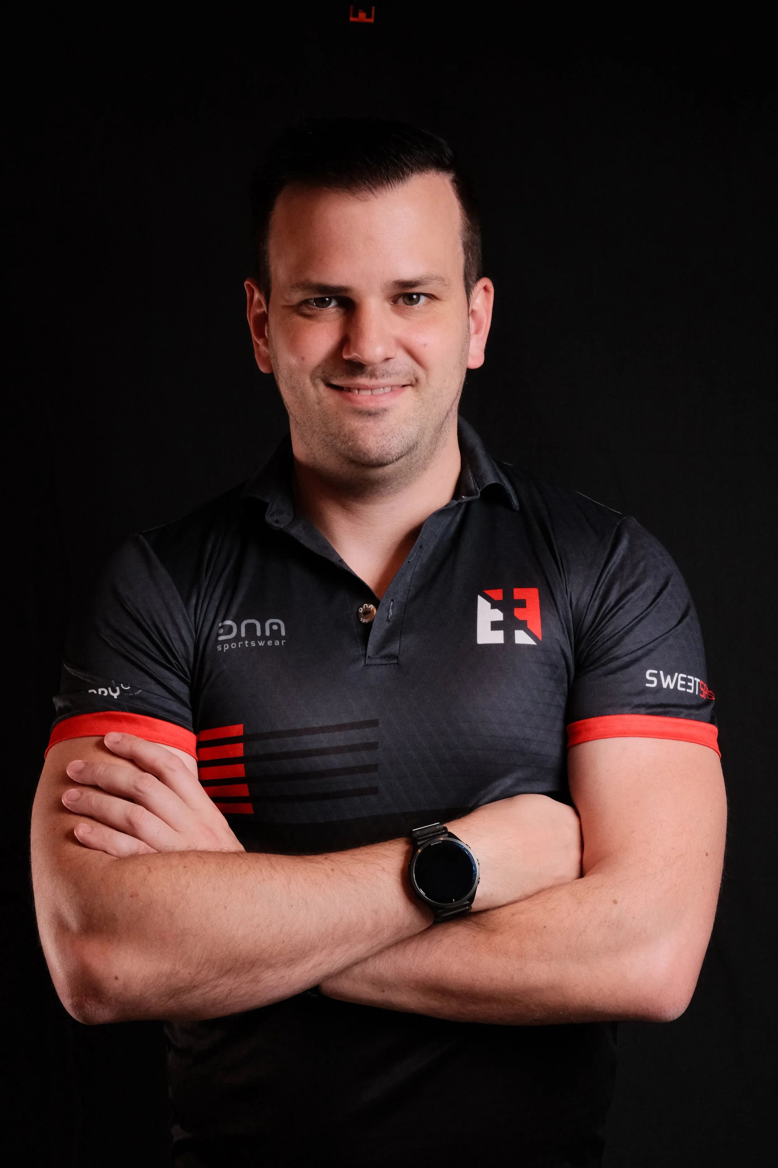 Ein Mann mit schwarzen Haaren, in sportlicher Kleidung, steht vor einem schwarzen Hintergrund, mit verschränkten Armen und einem freundlichen Lächeln.