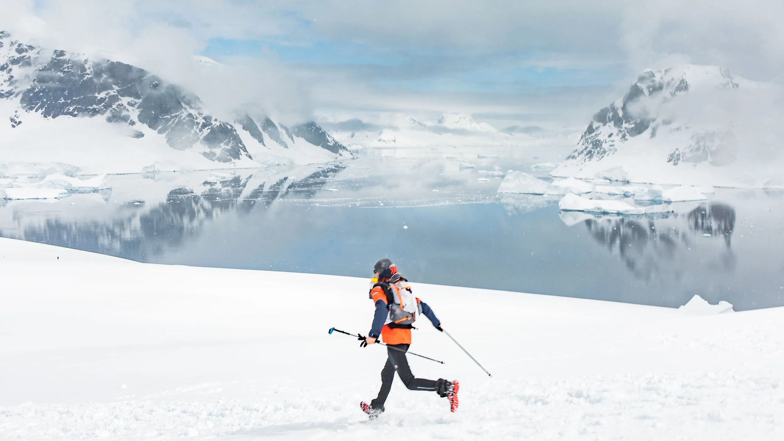 Ein Person in Winterkleidung und Stiefeln mit Schneeschuhen läuft durch eine verschneite Schneelandschaft in der arktischen Region, im Hintergrund schneebedeckte Berge und ein Eisfjord mit Eisschollen im Wasser.