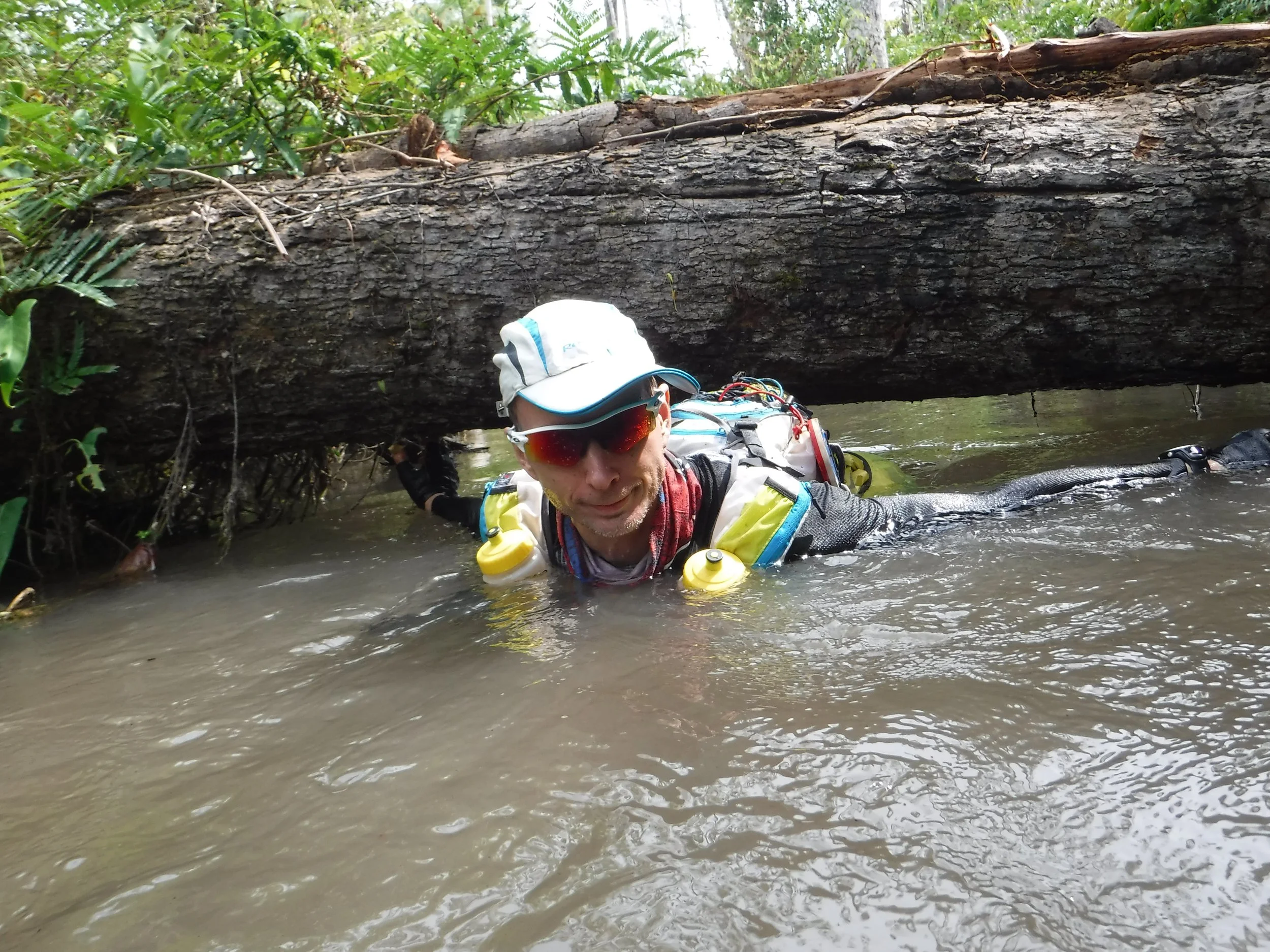 Mann trägt bei Flussüberquerung Schutzbrille, Hut und Rettungsweste, liegt im Wasser unter umgefallenen Baum, um den Fluss zu überwinden.