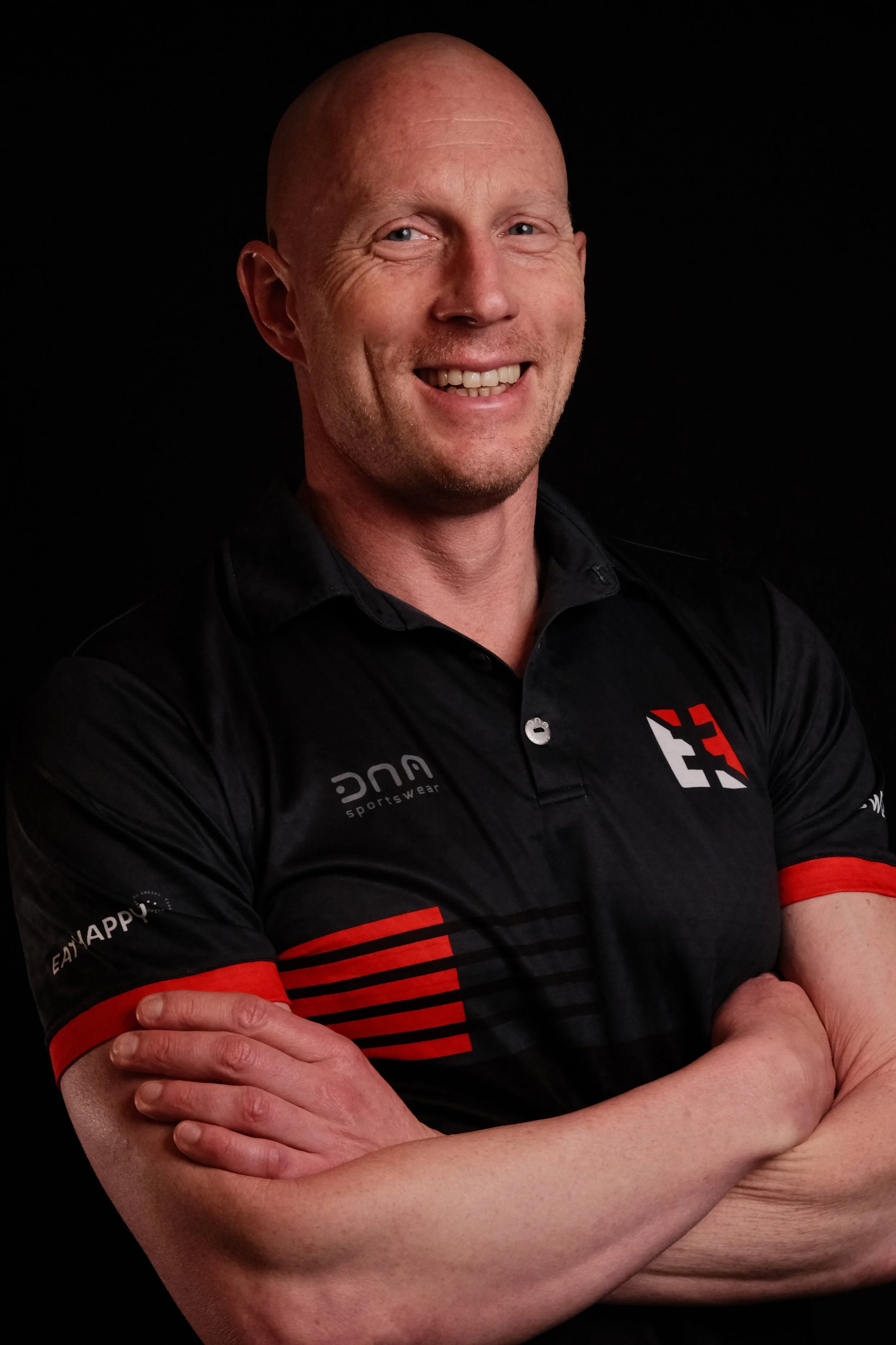 Ein Mann mit blauen Augen und Glatze trägt ein schwarzes Sporthemd mit roten Akzenten und lächelt freundlich. Er steht vor einem schwarzen Hintergrund.