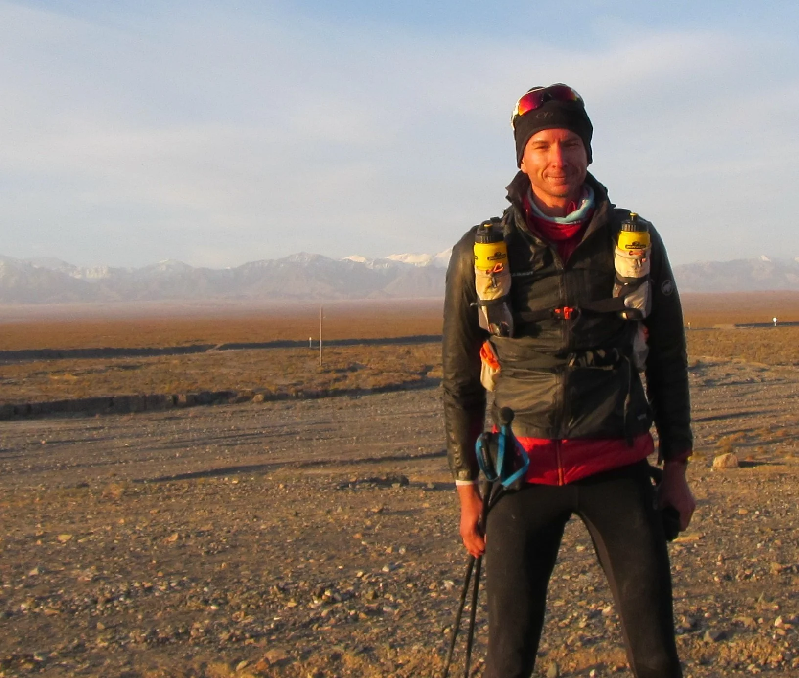 Ein Mann steht in einer verlassenen Wüste mit Bergen im Hintergrund, trägt Outdoor-Bekleidung und hat Wasserflaschen auf den Schultern.