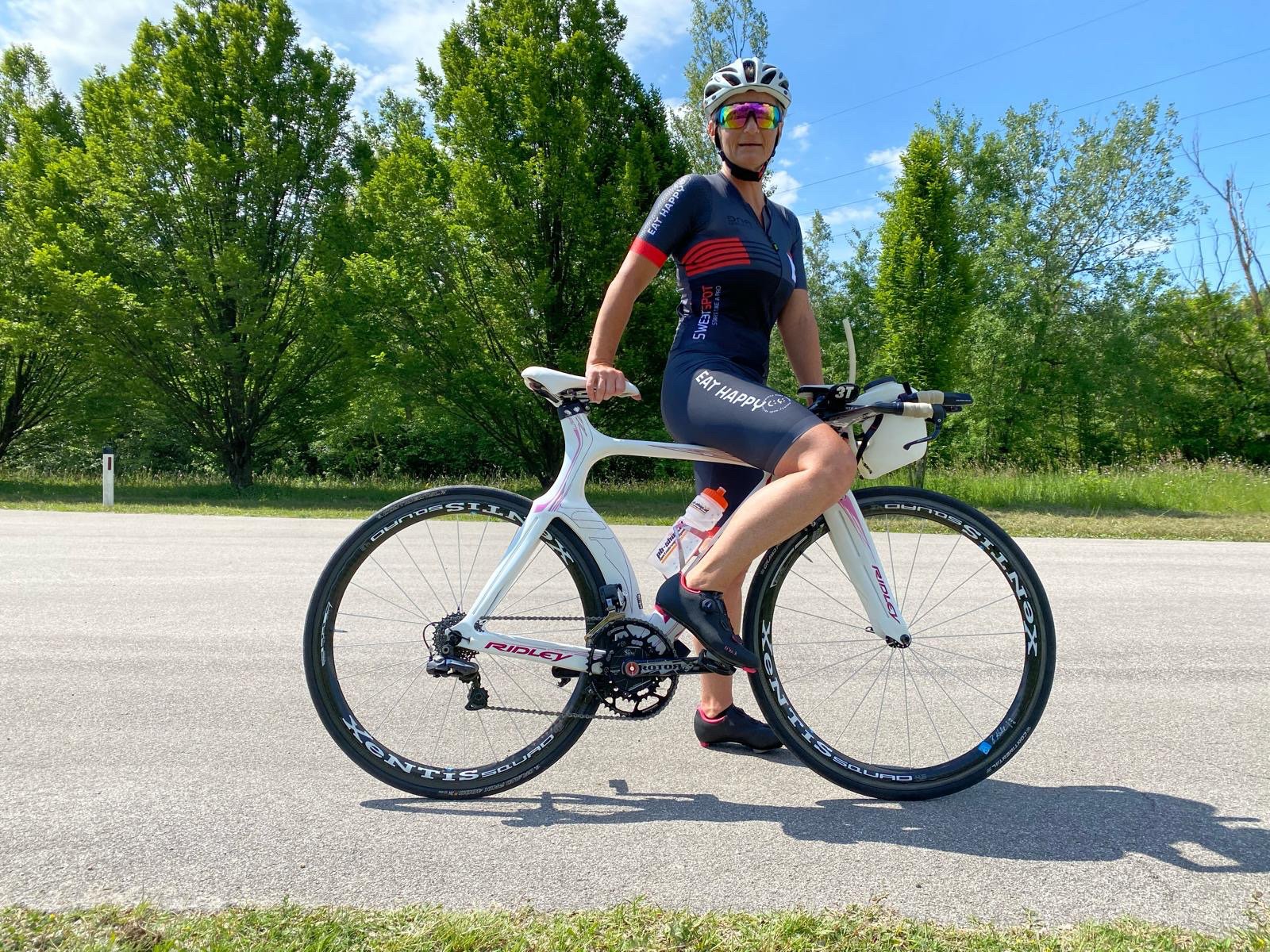 Frau in Radlertrikot mit Helm und Sonnenbrille auf Rennrad auf einer Straße, im Hintergrund Bäume und blauer Himmel.