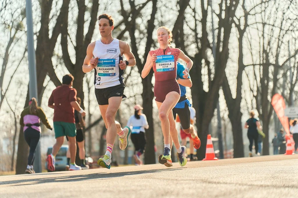 Zwei Läufer beim Straßenlauf, männlich und weiblich, mit Laufnummern, im Hintergrund weitere Teilnehmer, Bäume und orange Verkehrskegel, sonniger Tag.