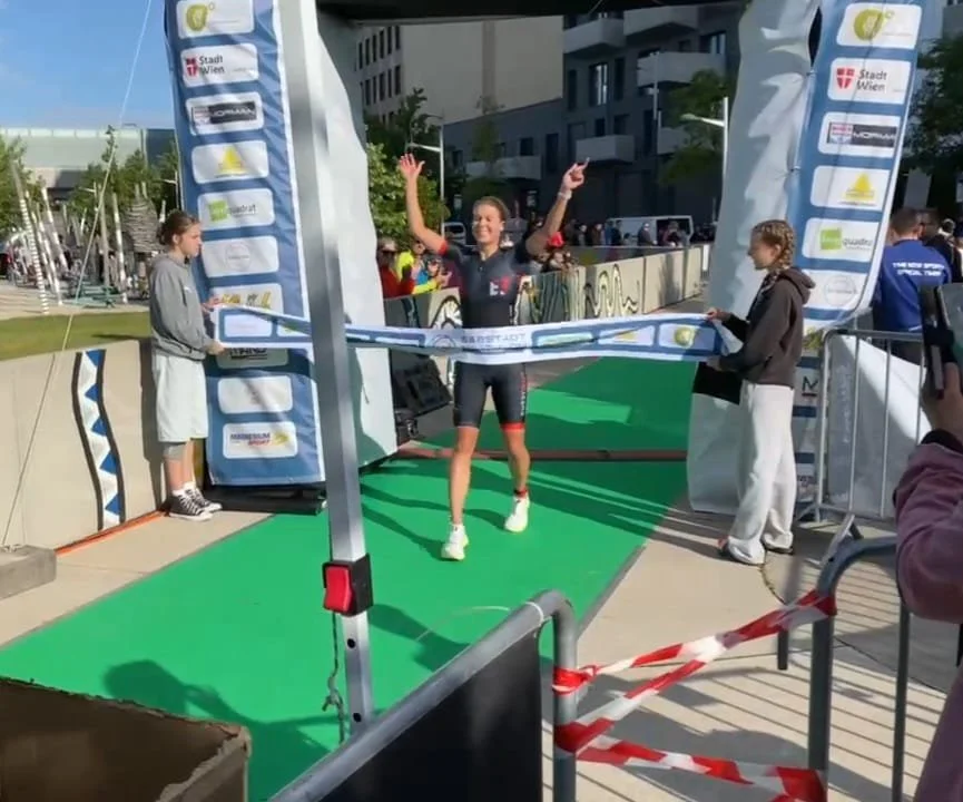Eine Frau beendet einen Triathlon, indem sie die Ziellinie überquert. Sie trägt einen schwarzen Triathlonanzug und hat die Arme in der Luft. Es ist ein Zielbogen mit mehreren Sponsorenlogos, und die Szene findet im Freien statt, vermutlich am Nachmit
