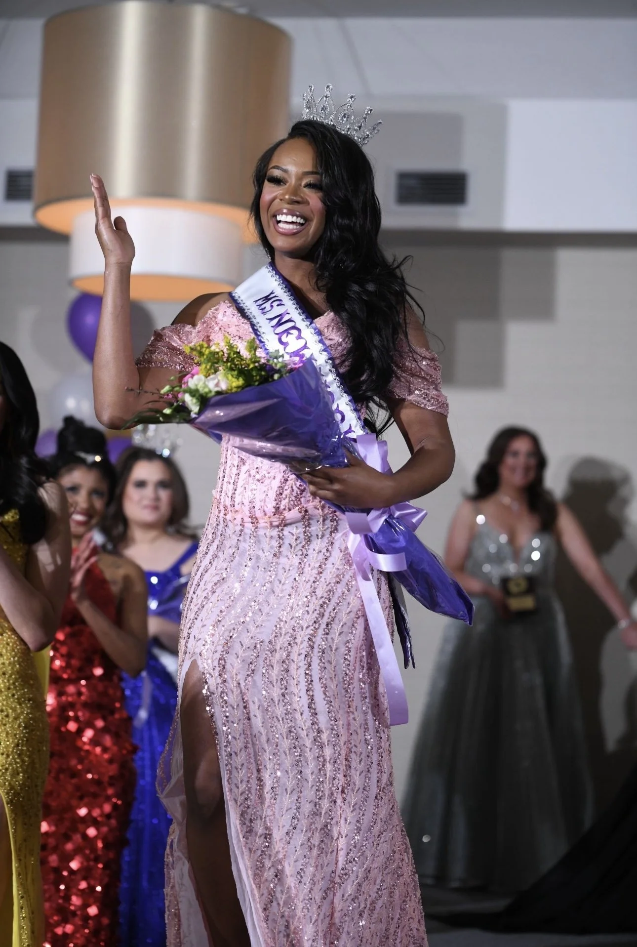 A woman wearing a crown and sash that reads 'Ms New York 2026' is holding a bouquet of flowers and smiling at a pageant. Miss New York. Ms New York Queenasia Rosario