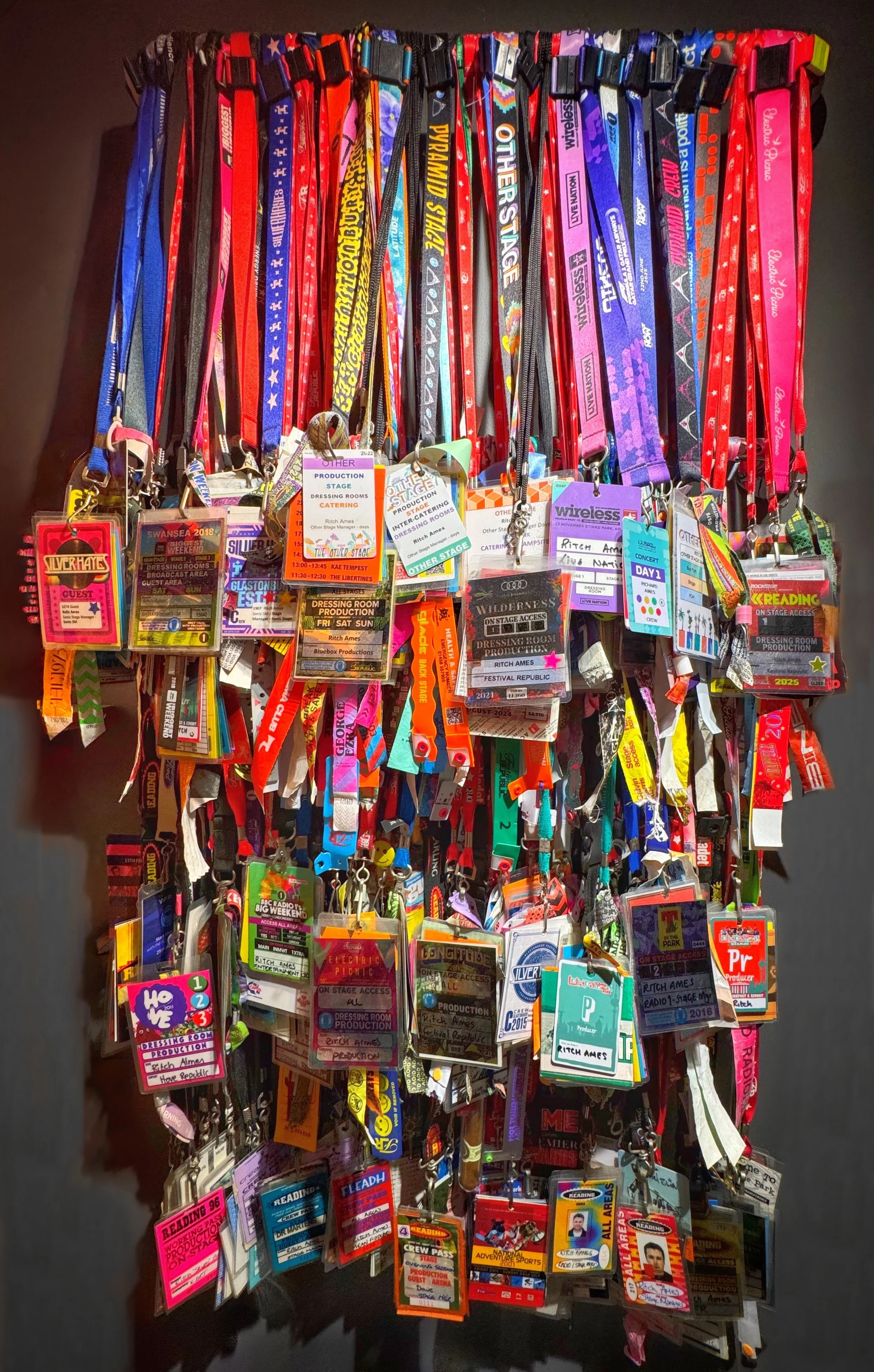 A large collection of colorful backstage pass and badge lanyards hanging on a wall, used for events, access, or identification showing Ritch Ames's stage management experience since 1992