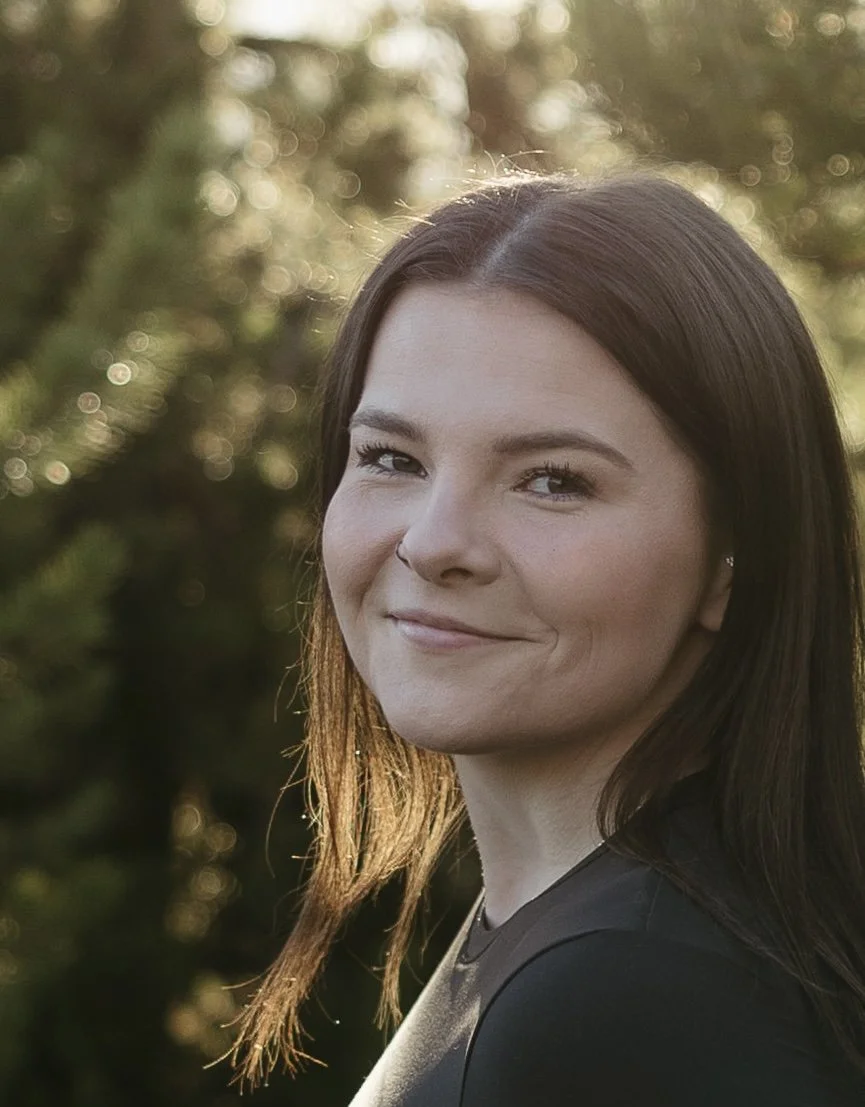 A woman with brown hair and a subtle smile, outdoors during sunset with blurred trees and sunlight in the background.