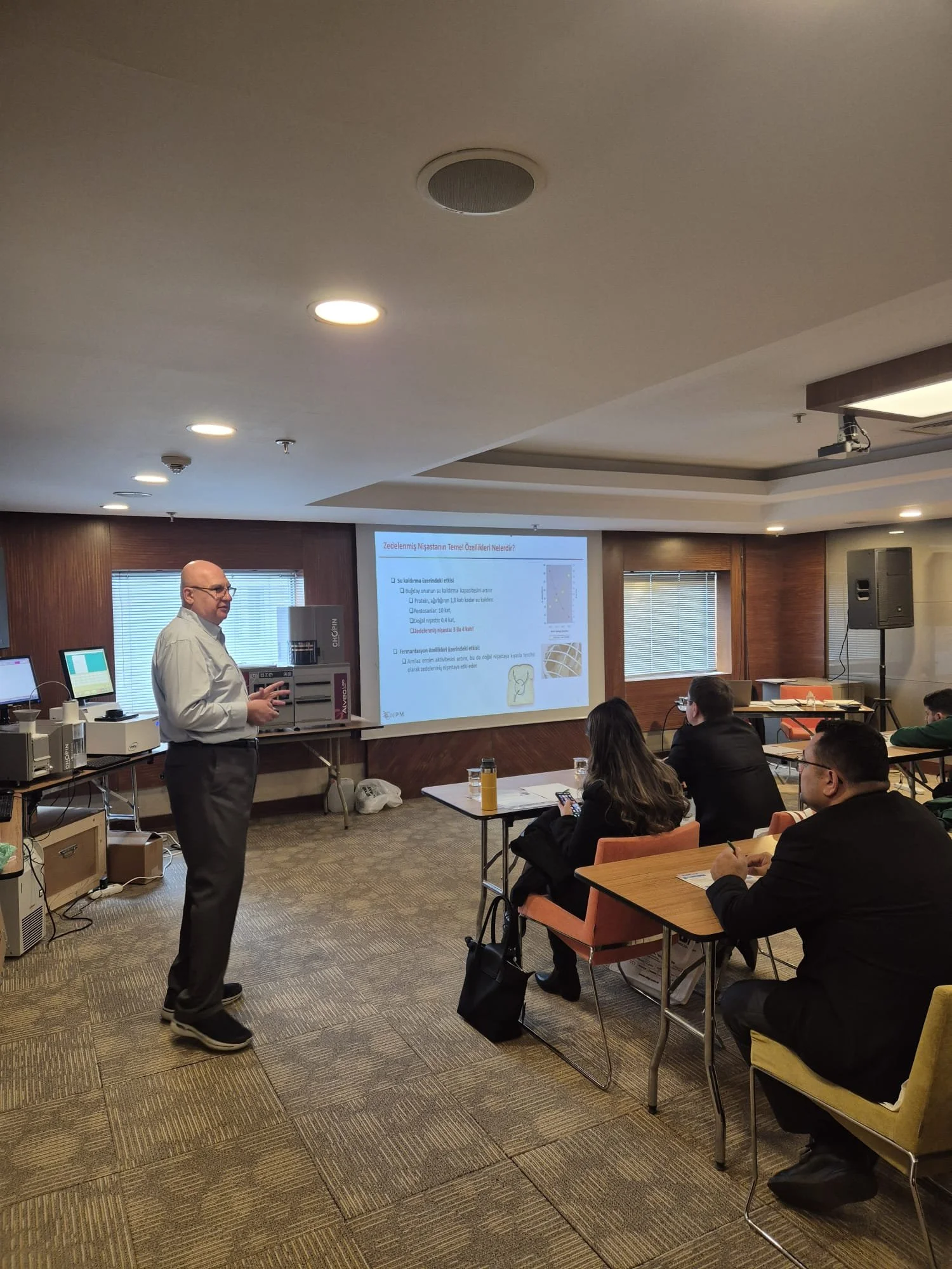 A man is giving a presentation in an office conference room with several people sitting at tables, a large screen displaying slides, and equipment on the side.