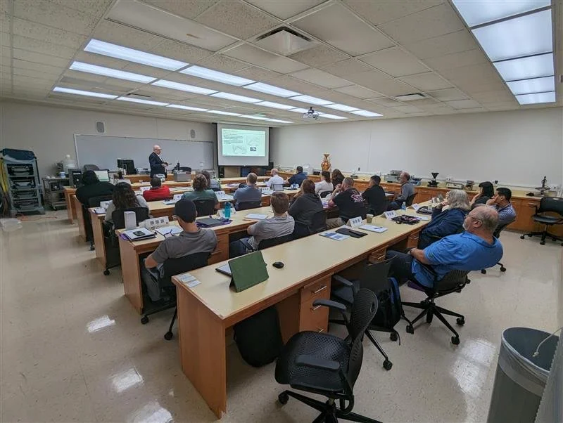 A classroom or conference room with a group of people attending a lecture or presentation. The presenter is at the front near a large whiteboard and a projection screen, with attendees seated at desks facing him.