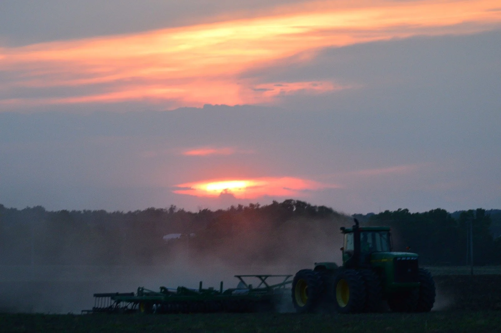 Farm tractor working in a field at sunset with a colorful sky and distant trees in the background.