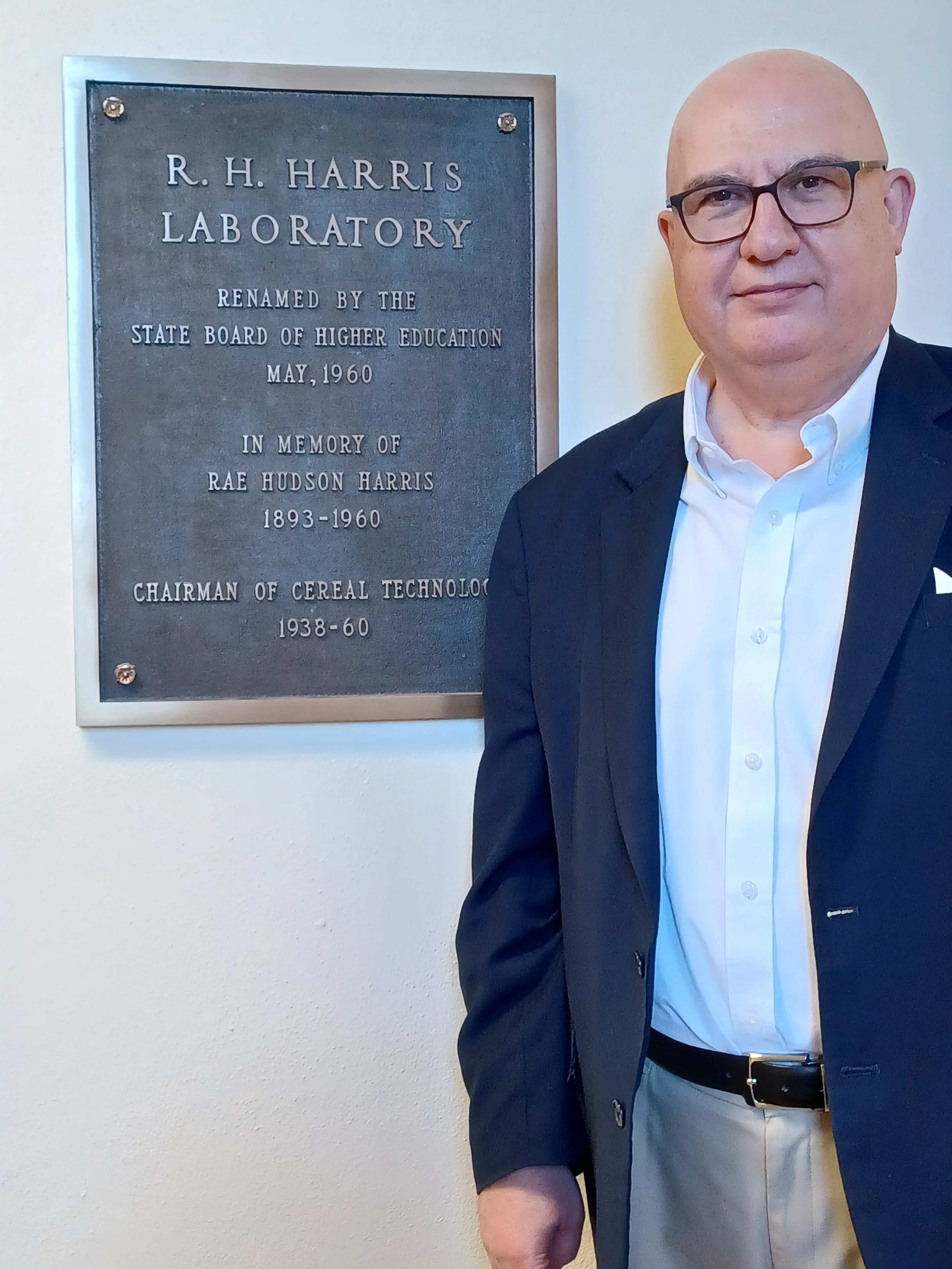 A man wearing glasses, a dark blazer, a white shirt, and light-colored pants standing next to a plaque that reads 'R. H. Harris Laboratory, Renamed by the State Board of Higher Education, May 1960, In Memory of Rae Hudson Harris 1893-1960, Chairman of Cereal Technology 1938-1960.'