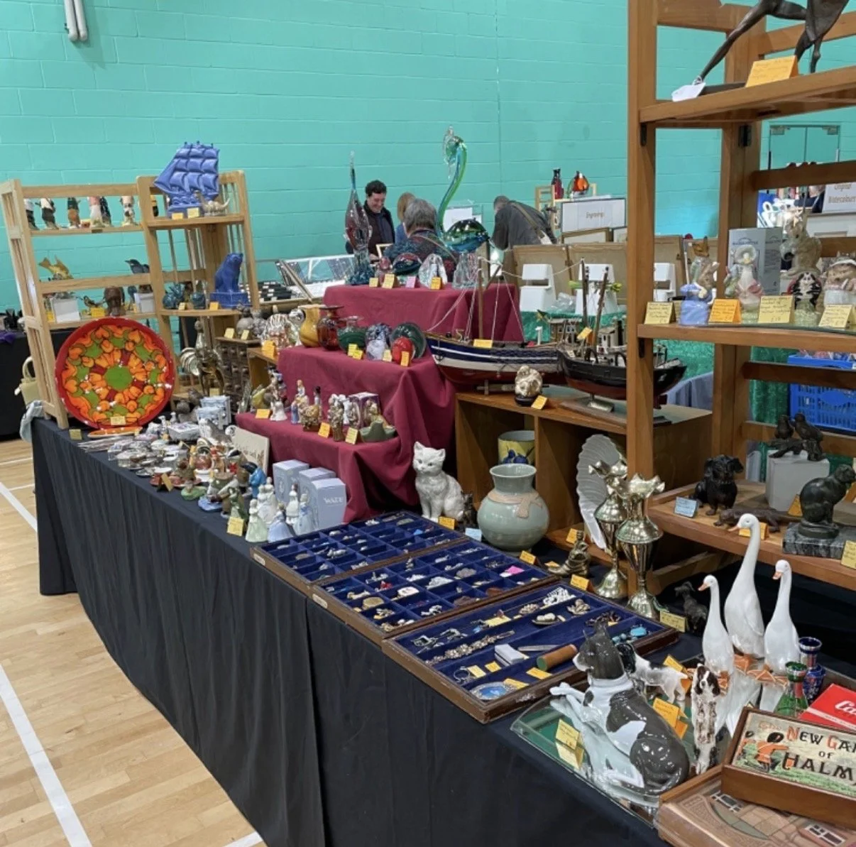 A display of various collectibles and decorative items at a craft fair, including figurines, glassware, pottery, jewelry, and model ships, arranged on tables covered with red and black cloths with a bright turquoise wall in the background.
