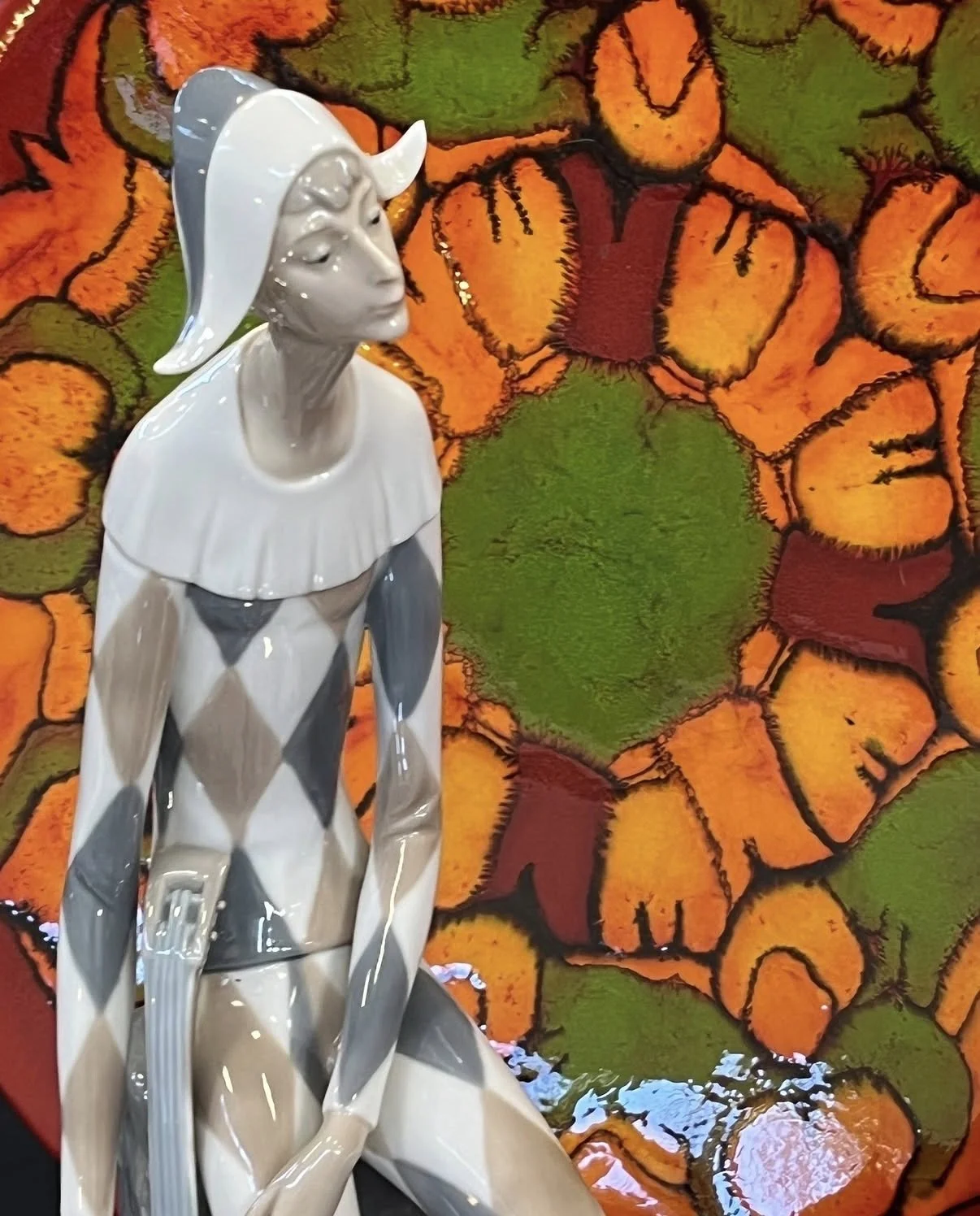 A white and gray ceramic sculpture of a woman with short hair, wearing a hat and a cape with a ruffled collar, is placed in front of a colorful, abstract floral mural with orange, green, and yellow hues.