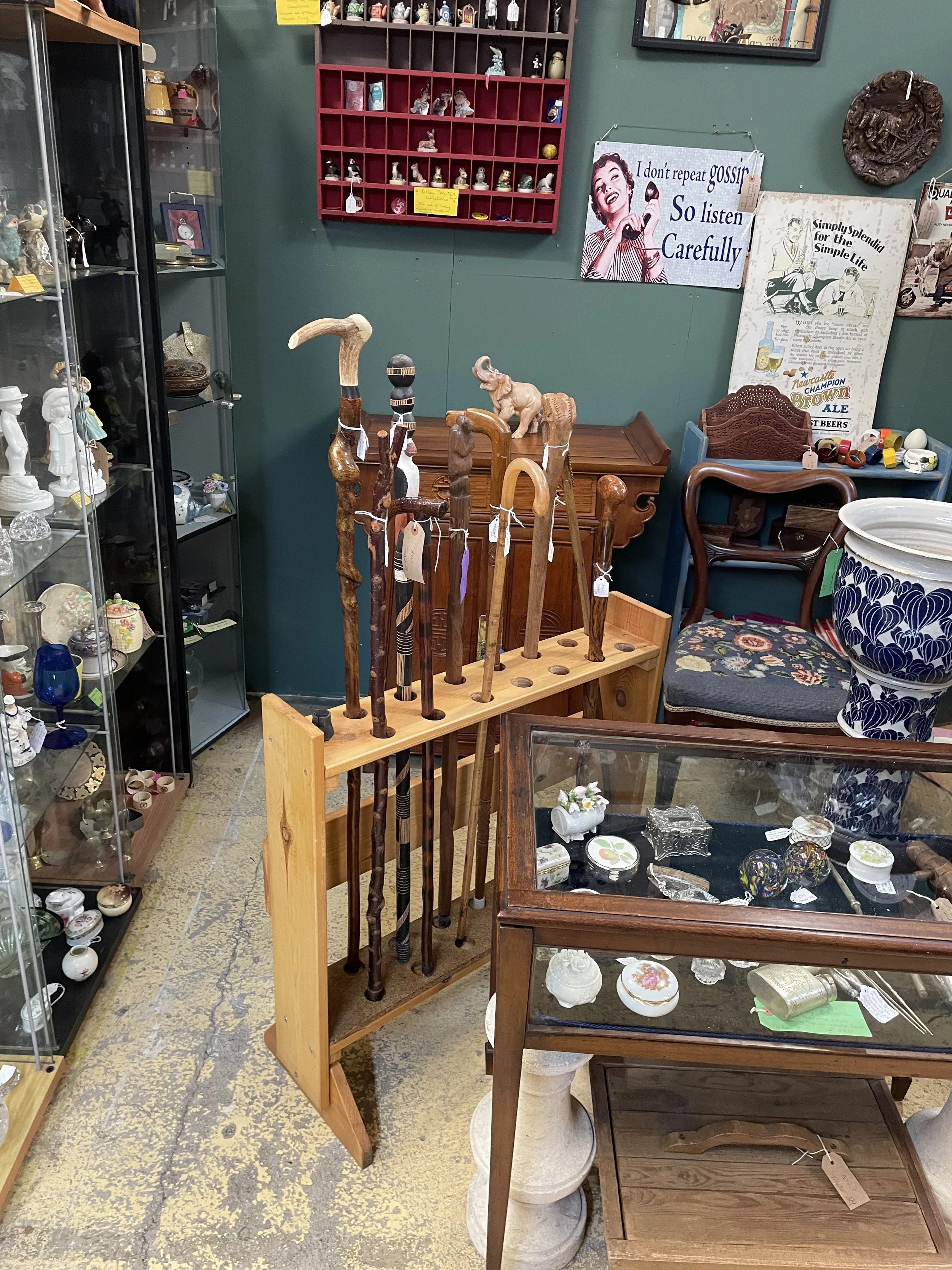 A display of wooden walking sticks with carved handles in a thrift store, with a glass display case filled with small decorative items and various antique furniture and signs on the walls.
