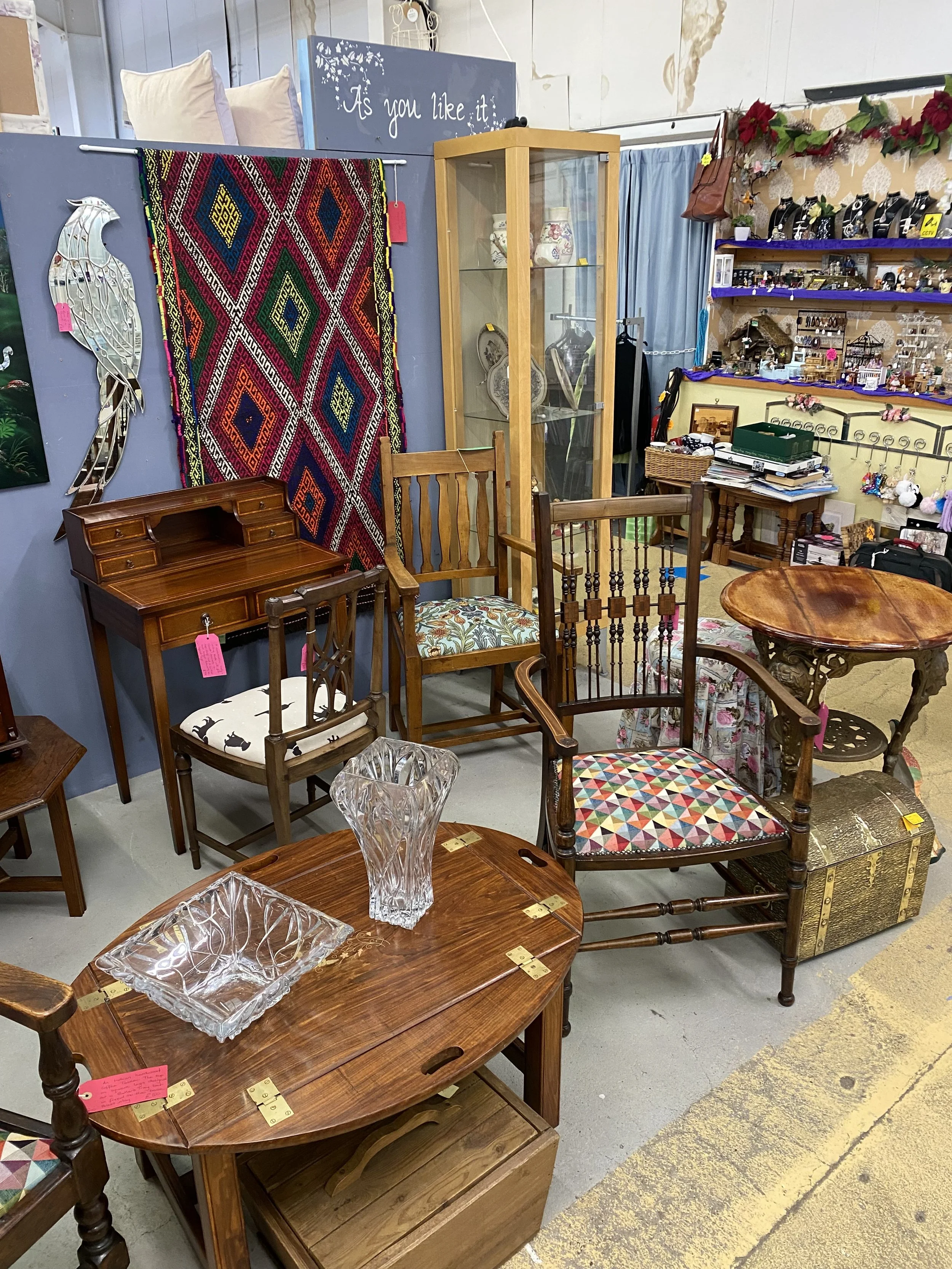 A collection of vintage furniture and decor items in an antique shop, including chairs, tables, glassware, a rug, and various trinkets.