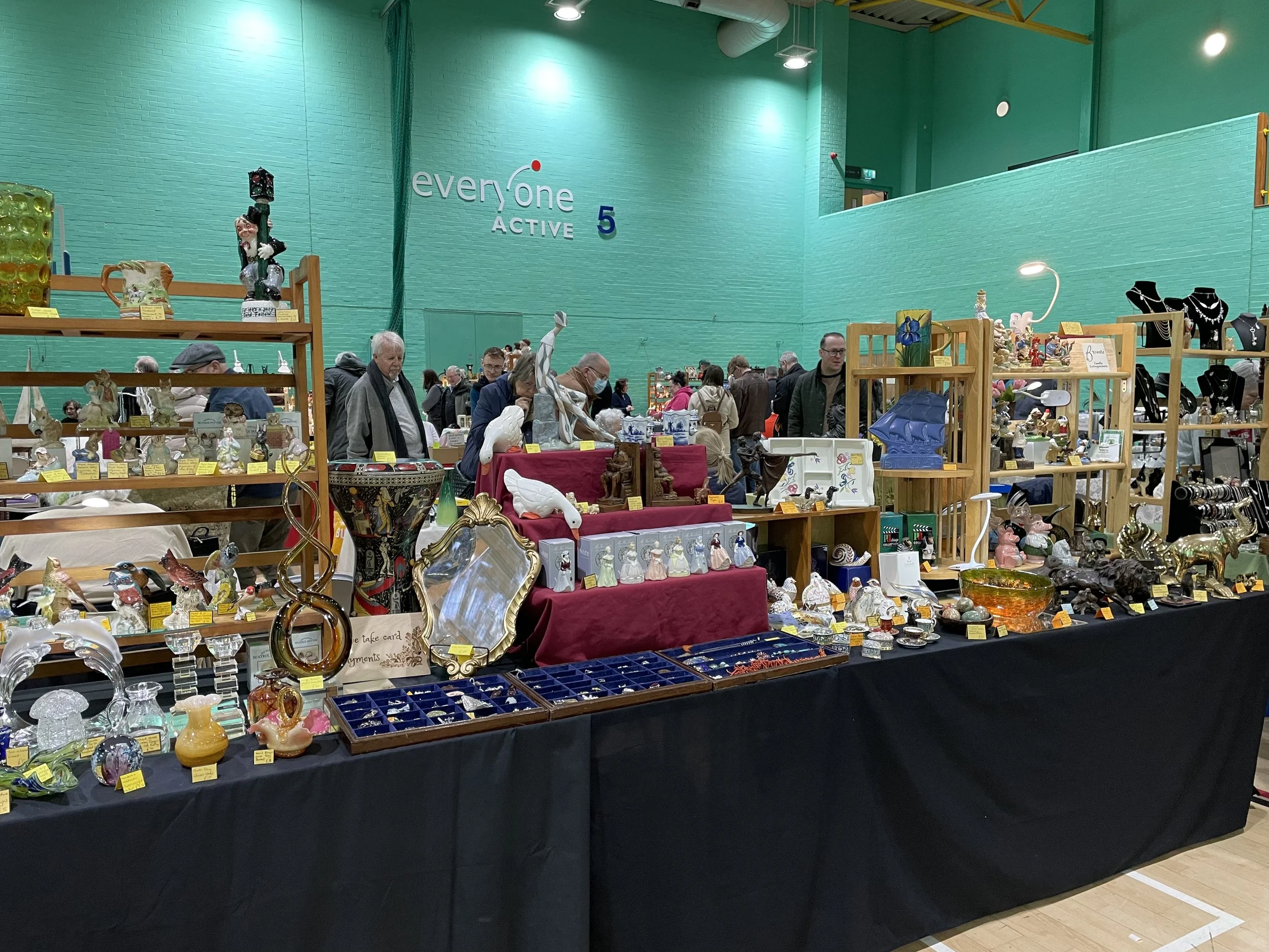 Indoor market or swap meet with tables and shelves filled with various collectibles, jewelry, and art pieces. Green walls and several shoppers browsing the items.