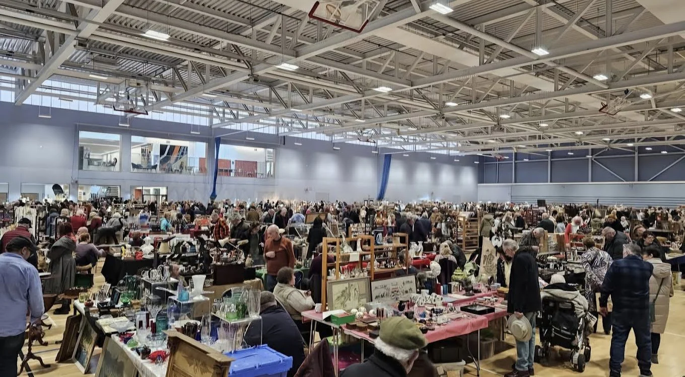 A large indoor flea market or indoor market with many tables and booths filled with various items like glassware, art, and collectibles, crowded with shoppers browsing and shopping.