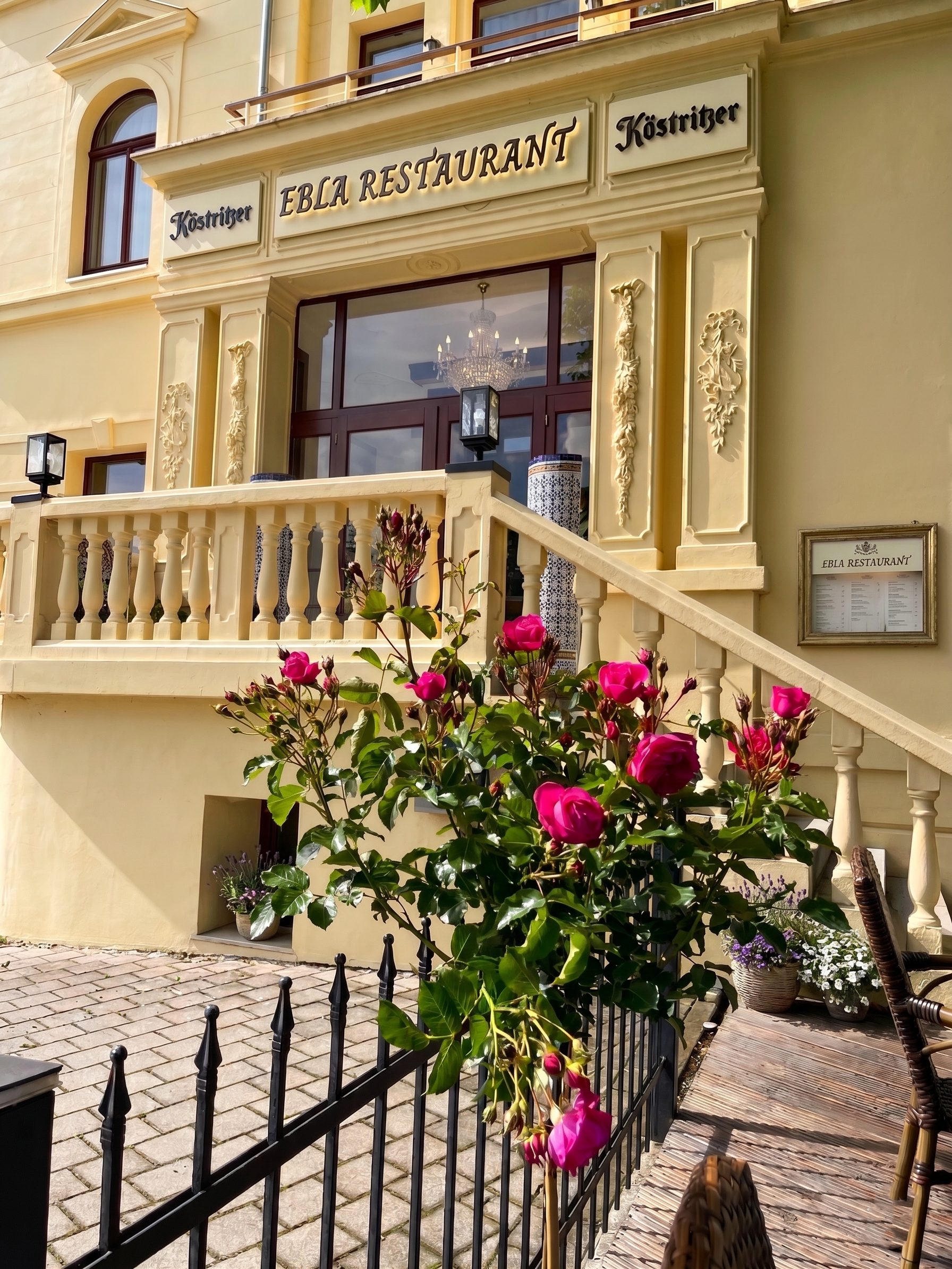 Vorderansicht eines gelben Restaurants namens 'Ebla Restaurant Köstritzer' mit Balkon, blühenden rosa Rosen im Vordergrund und einem Kaktus sowie Blumen in Körben vor der Terrasse.