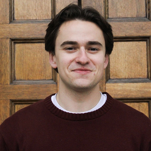 A man with short, dark hair, wearing a red sweater and white undershirt, smiling and standing in front of a brown, wood door.