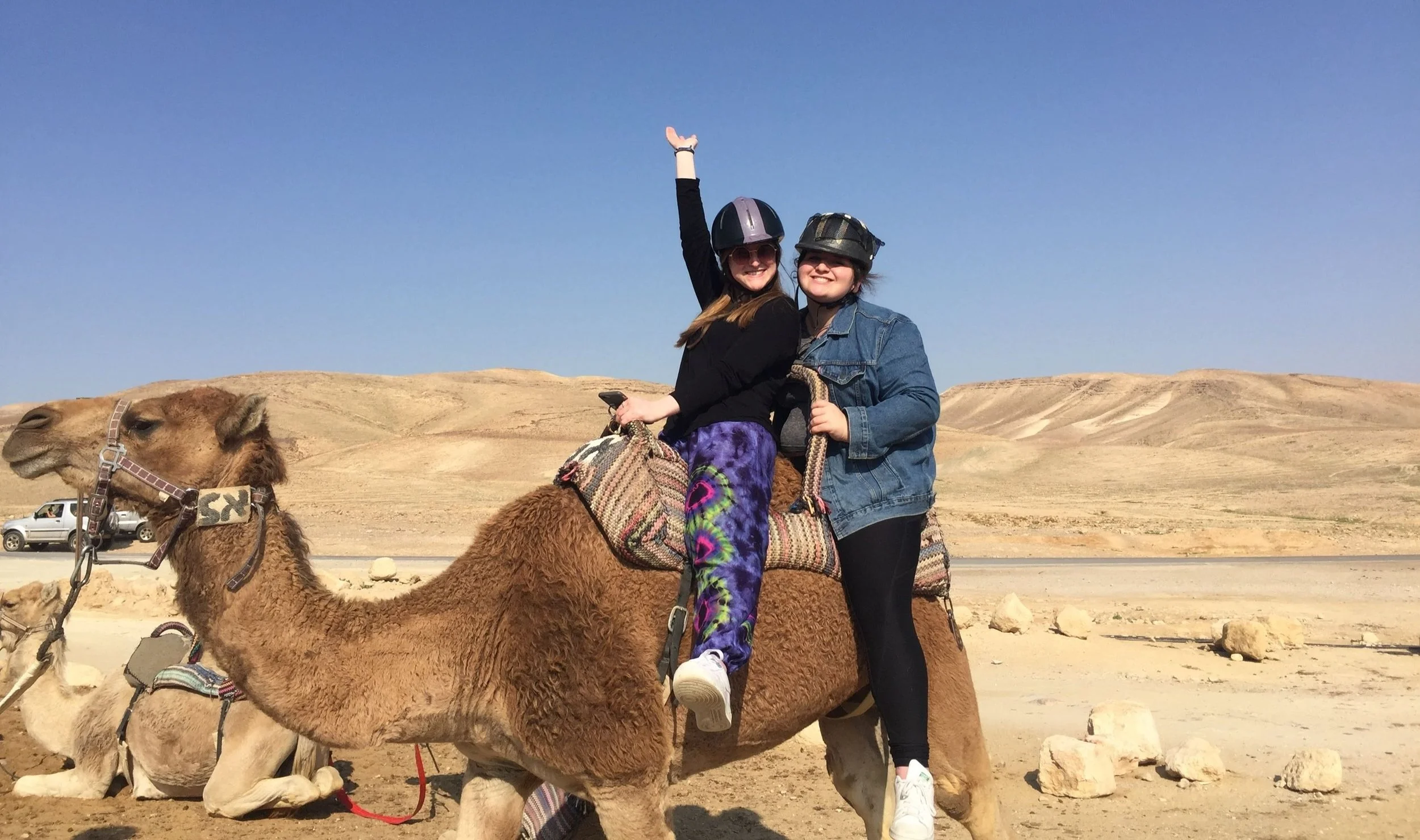 Two women riding camels in a desert landscape, smiling and wearing helmets.