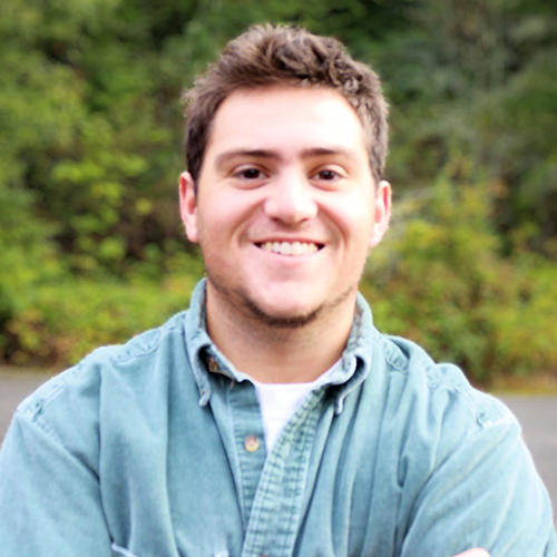 A man with short brown hair, wearing a blue shirt, smiling and standing outdoors with bushes behind him.