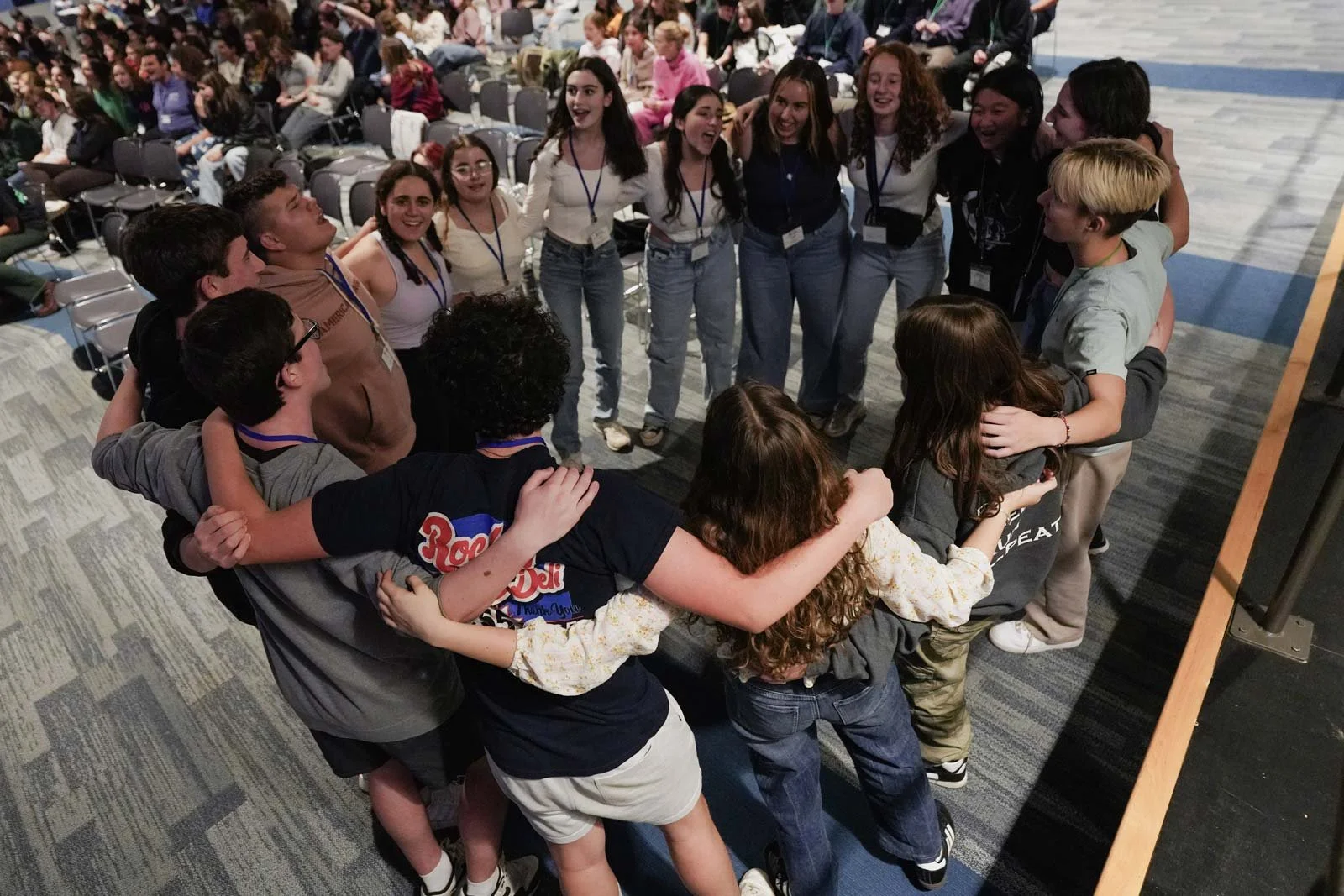 A group of young people standing in a circle with their arms around each other, smiling and laughing, in a large indoor conference or auditorium setting.