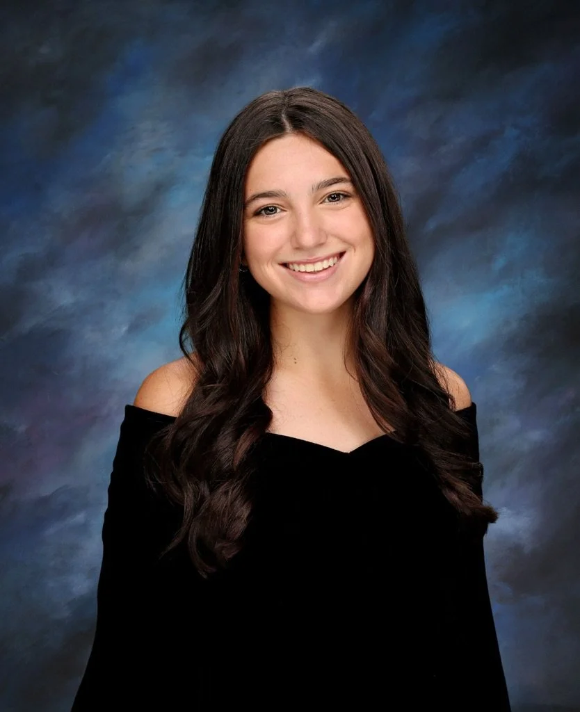 Young woman with long dark hair smiling, wearing an off-the-shoulder black top, against a blue and gray studio background.