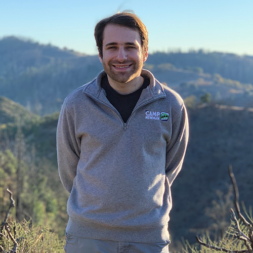A man with brown hair, wearing a gray sweatshirt, standing with his arms behind his back, outdoors with moutains behind him.