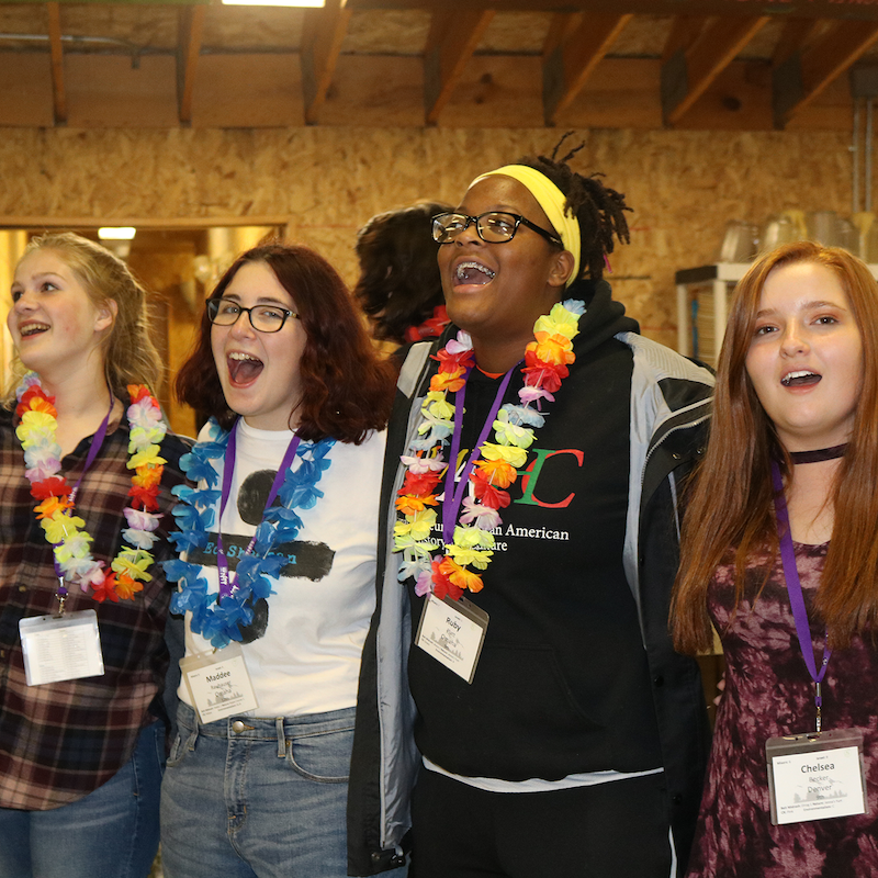 Group of young people laughing and smiling, wearing colorful leis, indoor setting with wooden walls, casual attire, some with glasses and name tags.