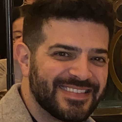A close-up of a man's face with dark hair and facial hair, smiling.