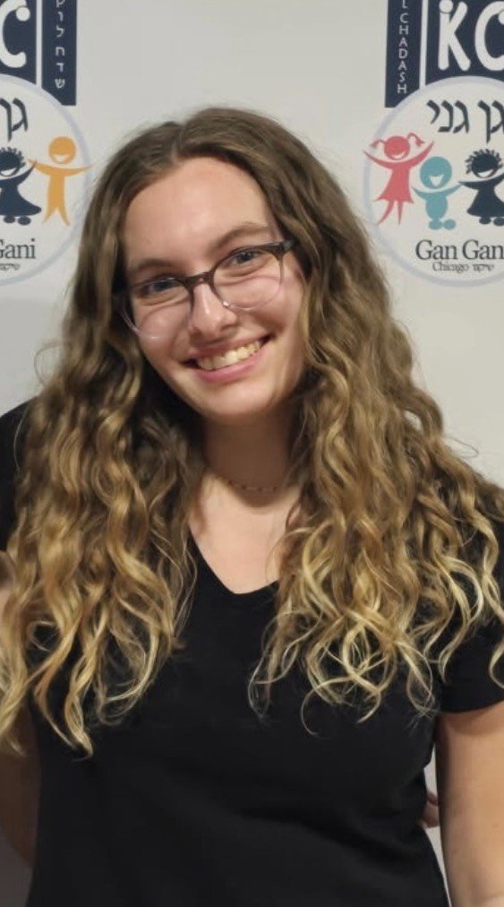 A young woman with long, curly blonde hair wearing glasses and a black shirt, smiling at the camera, standing in front of a white backdrop with colorful logos.