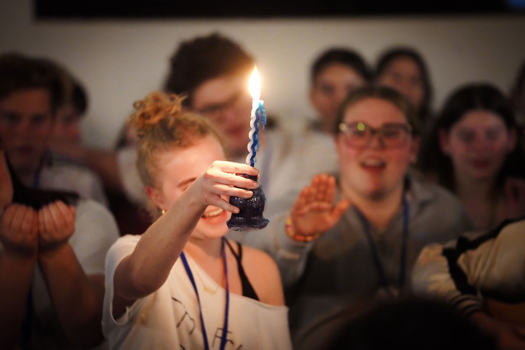 Group of people celebrating indoors with a birthday cake and a lit candle, smiling and cheering.