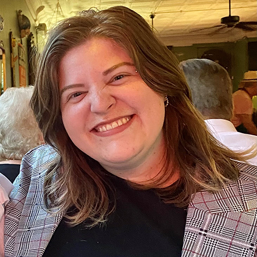 A woman with shoulder-length hair smiling, wearing a dark shirt and black and white jacket.