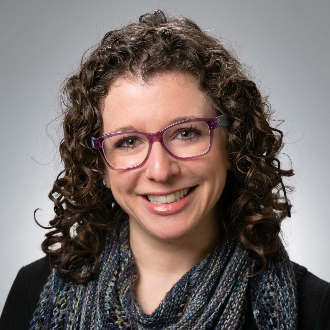 A woman with curly brown hair wearing purple glasses and a colorful scarf, smiling against a plain background.