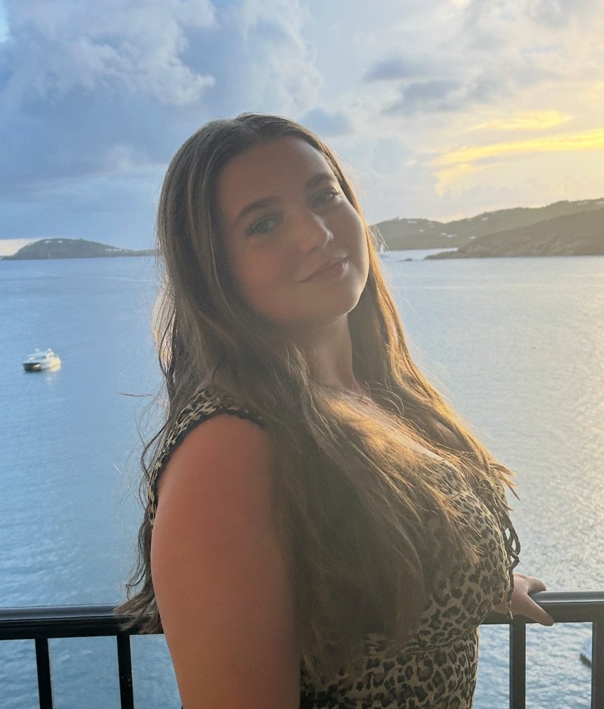 A woman with long brown hair standing on a balcony overlooking a body of water with islands in the distance during sunset.