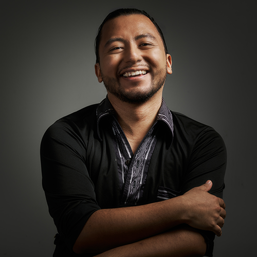 A man standing with his arms crossed, wearing a dark shirt and smiling, standing in front of a solid dark background.