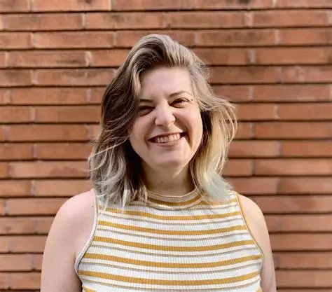 A woman with shoulder-length hair smiling in front of a brick wall, wearing a sleeveless striped top.