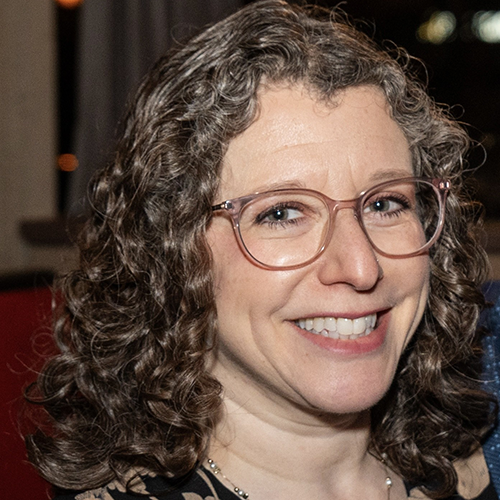 A woman with curly brown hair wearing glasses, smiling.