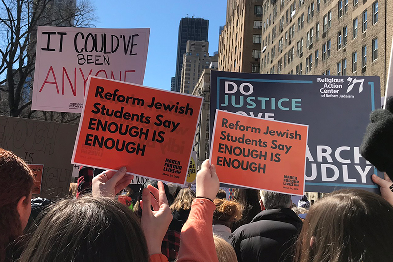 Crowd of people protesting with signs, including one that reads "Reform Jewish Students Say ENOUGH IS ENOUGH" and another with the words "DO JUSTICE LOVE". Tall buildings and a clear sky in the background.